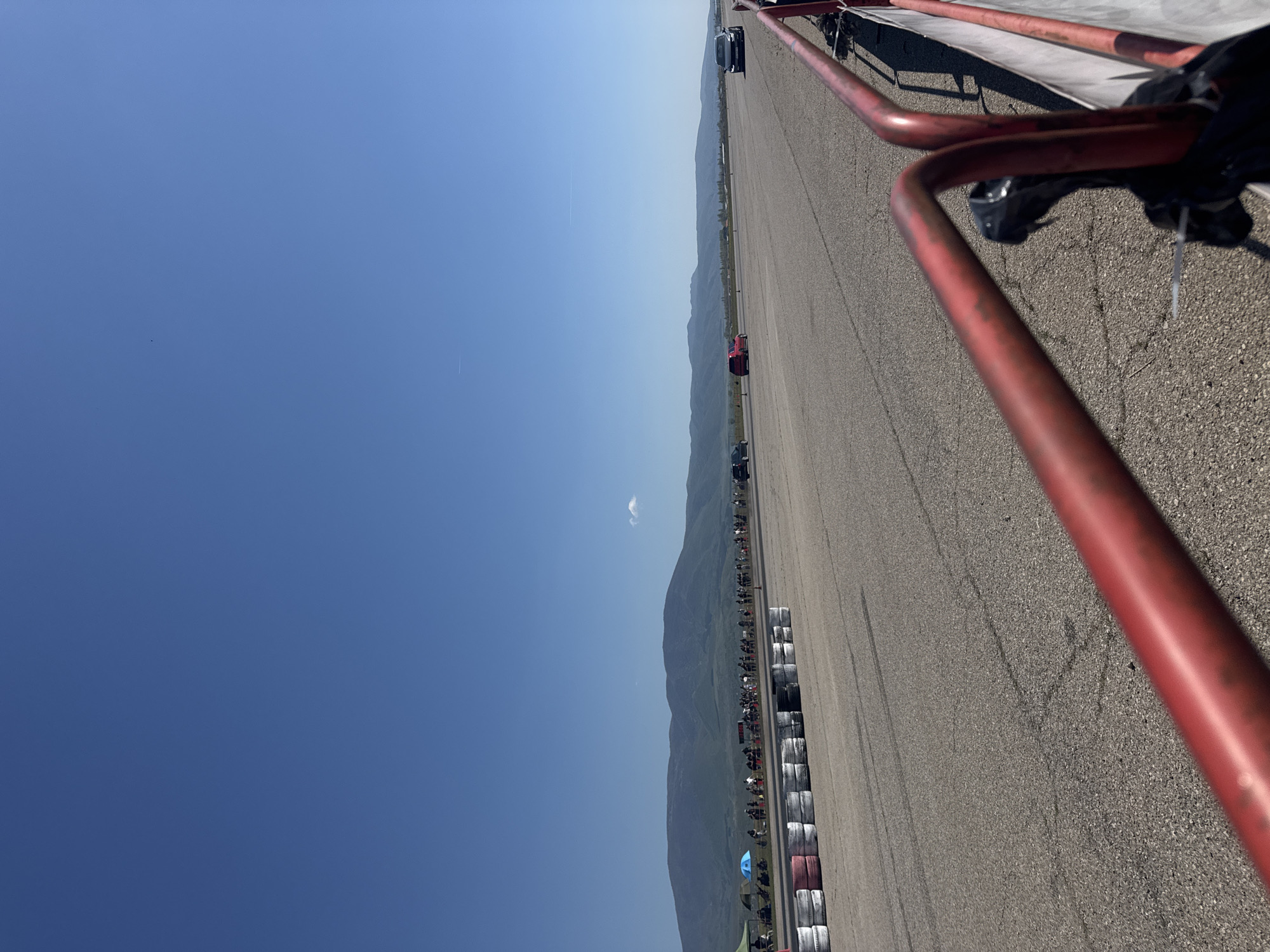 The image shows a long, flat asphalt racetrack stretching into the distance, with a line of tire barriers running along one side. Beyond the track and the tire barriers are low-lying hills or mountains under a clear blue sky. Several vehicles, including cars, can be seen on the track at varying distances. A red-painted metal barrier is visible in the foreground on the right side of the image. Many people are present alongside the tire barriers. The location is Sliven, Bulgaria.