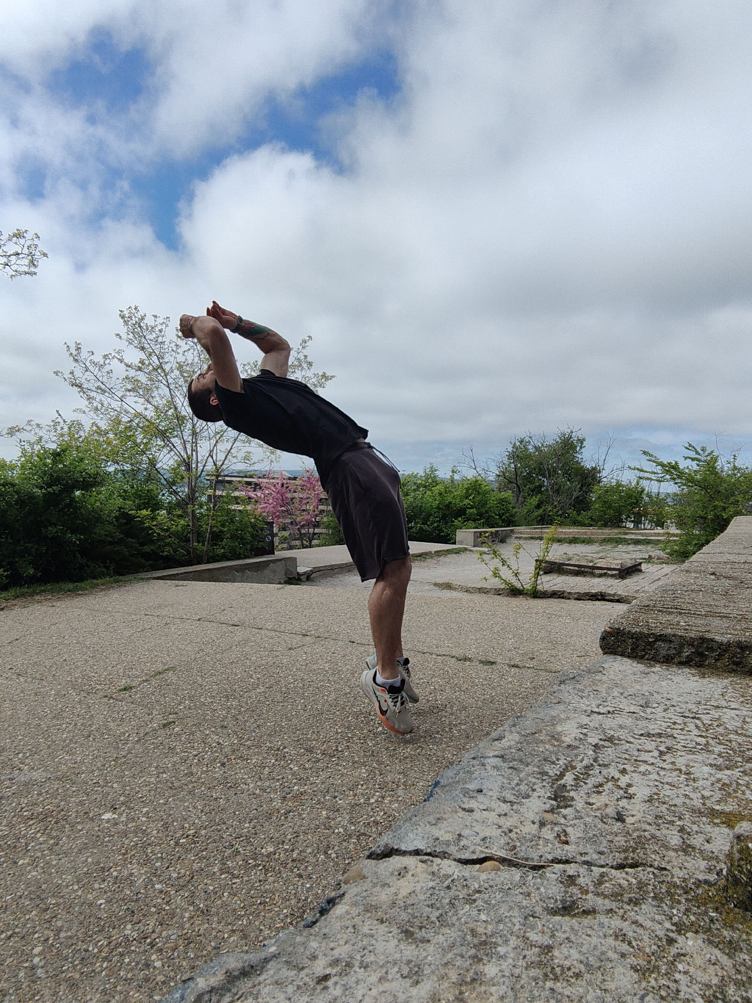 A man is captured mid-backbend in an outdoor setting in Varna, Bulgaria. He is wearing a black t-shirt, dark shorts, and running shoes, with a tattoo visible on his arm. His arms are raised above his head, bent at the elbows, as he performs the acrobatic move.
The backdrop includes a cracked concrete surface and low concrete structures, possibly remnants of a park or recreational area. Green bushes and trees are visible behind the concrete, and a glimpse of the sea is seen in the distance. The sky is overcast with a mix of blue and grey clouds.