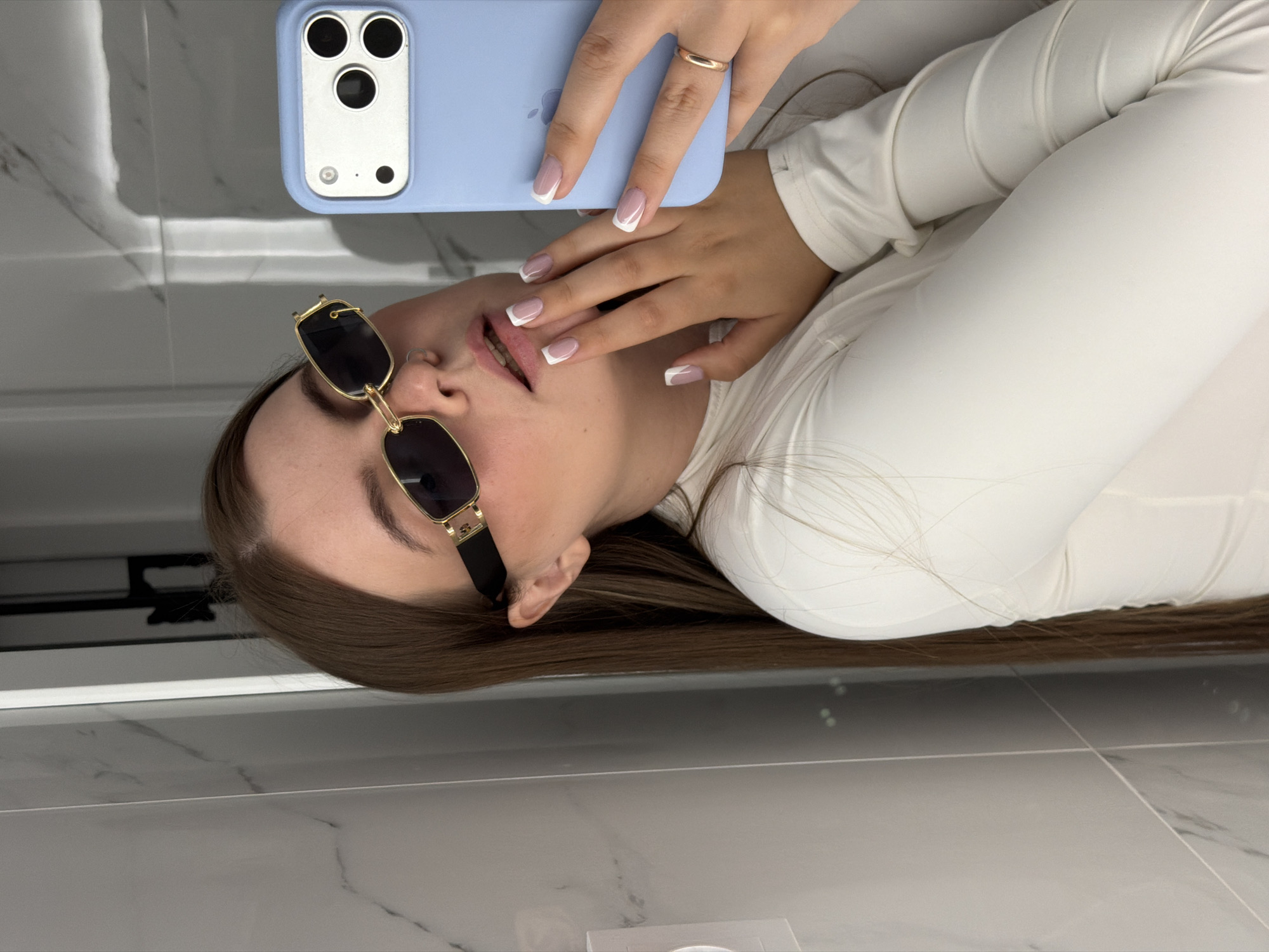 A person with brown hair and sunglasses is taking a selfie in front of a gray marble tiled background. The person is wearing a white long-sleeved shirt and holding a blue iPhone with three camera lenses. The person has manicured nails with white tips. The location is not apparent from the image.