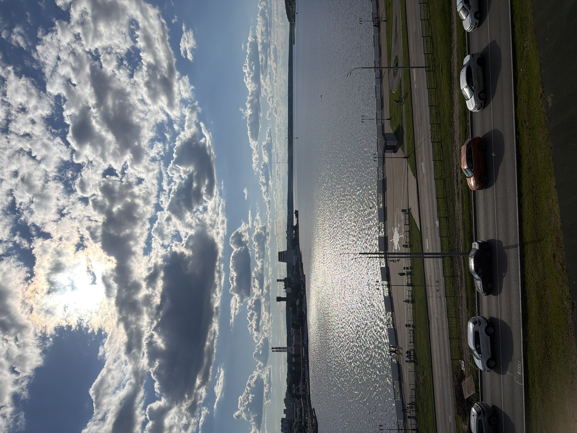 The image, taken from an elevated perspective, captures a wide panoramic view of a city waterfront under a partially cloudy sky. The bright sun is visible behind clouds in the upper left, casting a strong glare that reflects vibrantly on the expansive body of water, likely a river or reservoir, stretching across the middle of the frame.
In the distance, on the far bank of the water, a cityscape is visible, featuring several buildings, including what appear to be industrial structures or tall chimneys, indicating an urban or industrial area.
In the foreground, a multi-lane road runs parallel to the water. Several cars are present on the road, with five distinct vehicles visible: a dark grey/black car, two white cars, an orange/brown car, and a light grey/silver car. Their long shadows suggest a relatively low sun angle, indicating it is likely late morning or afternoon. Between the road and the water, a paved pedestrian path or embankment is visible, bordered by railings and interspersed with streetlights. A few small figures are discernible on this path, suggesting people are walking or cycling, with one person on a bicycle faintly visible. Grass strips line parts of the road and embankment.
The scene depicts a sunny day with scattered clouds. No discernible text is visible in the image. The overall impression is one of everyday urban life by a significant body of water, possibly the Izhevsk Pond in Izhevsk, Russia.