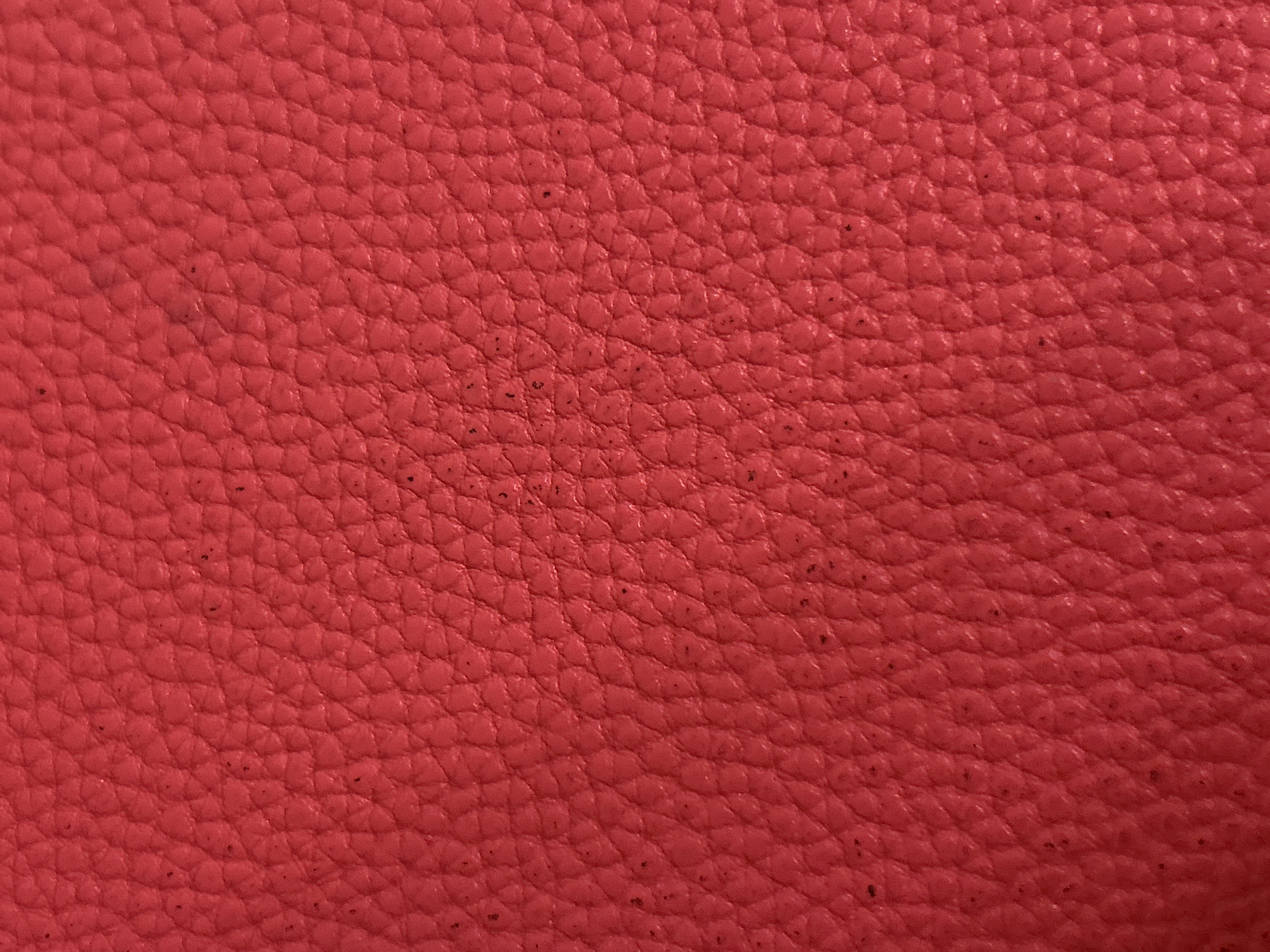 The image shows a close-up, textured surface of red leather. The leather has a fine-grained pattern with many small, irregular raised bumps. There are also some very small, dark specks scattered across the surface, which could be dirt or imperfections in the material. The lighting appears to be even, highlighting the texture of the leather without casting harsh shadows. The color is a vibrant, saturated red. There is no discernible information about the location within Moscow, Russia, nor are there any people, objects, actions, or interactions depicted.