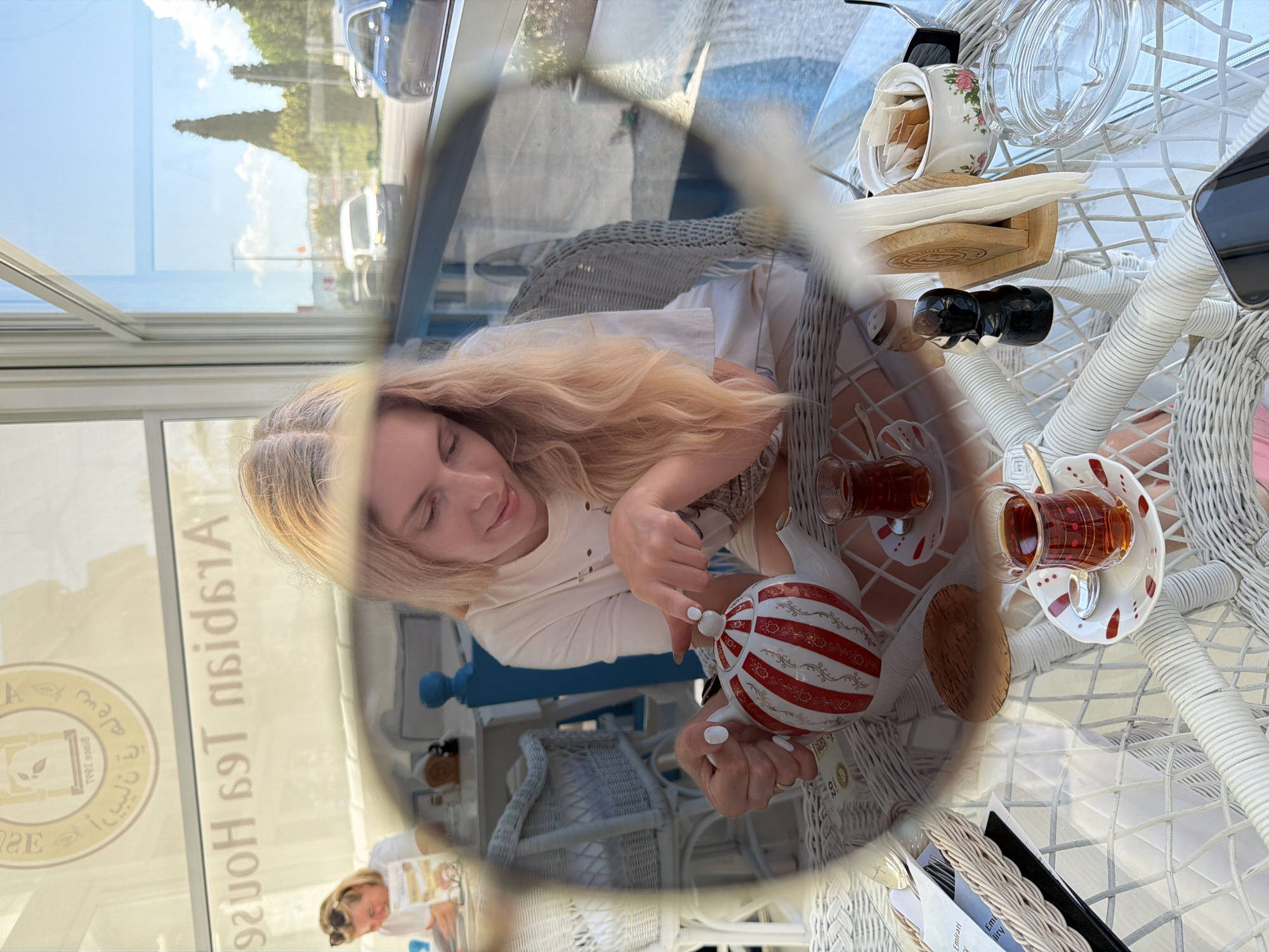 A fair-skinned, blonde-haired female in a white shirt is sitting at a white wicker table. She is interacting with a striped teapot near two tea cups on matching saucers. The table also holds a sugar bowl with packets, small wooden box, a small ceramic container with a black lid, a menu in a wicker holder, and a cell phone. The scene is inside a building with a window displaying the exterior and reflection of the interior with the logo for "Arslan Tea House" visible in the reflection. Podgorica, Montenegro, is presumed as the city location.