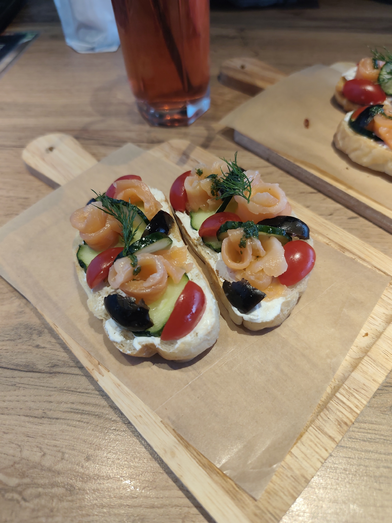 Two open-faced sandwiches (likely bruschetta or similar) are presented on a small, wooden cutting board lined with brown parchment paper. The sandwiches are topped with cream cheese, slices of cucumber, halved cherry tomatoes, black olives, and rolled smoked salmon, garnished with dill. In the background, there is a tall glass of dark pink liquid (possibly iced tea or juice) and a second cutting board with another sandwich. The scene is set on a wooden table. There is no verifiable information to confirm if the location is in the city of Chaikovsky, Russia.