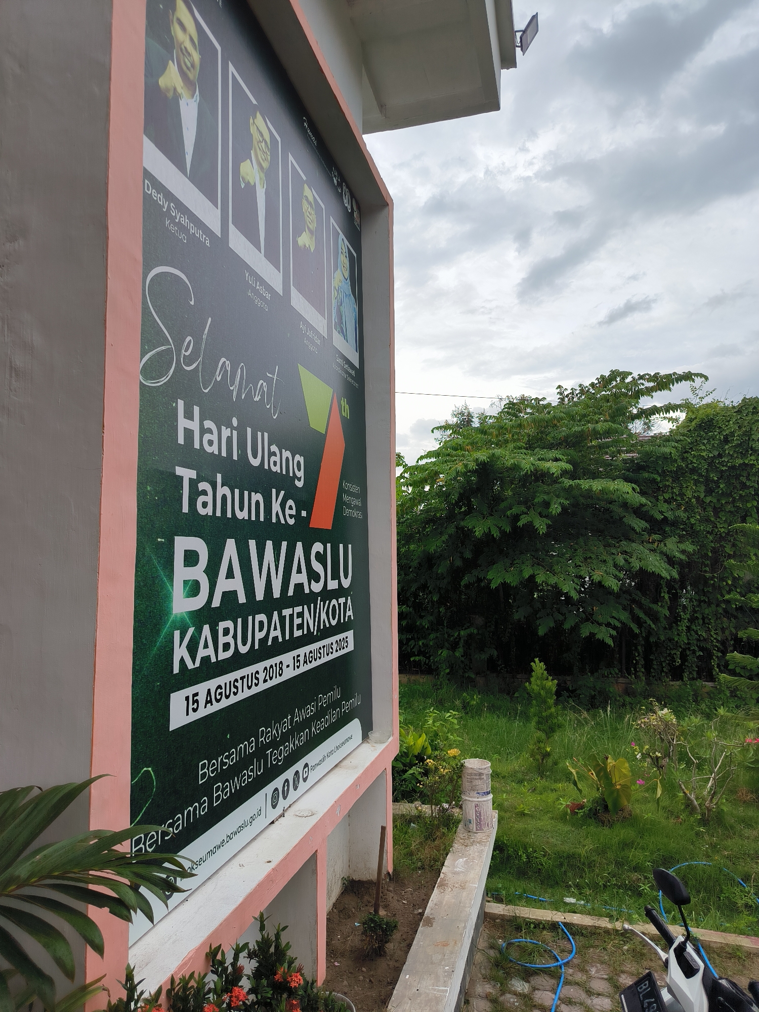 A large, vertical green banner celebrating the anniversary of BAWASLU KABUPATEN/KOTA (Regency/City Election Supervisory Agency) is prominently displayed on the light pink exterior wall of a building in Lhokseumawe, Indonesia. The banner reads "Selamat Hari Ulang Tahun Ke-7th BAWASLU KABUPATEN/KOTA" with the dates "15 AGUSTUS 2018 - 15 AGUSTUS 2025," indicating a 7th anniversary celebration within that period.
The top part of the banner features black-and-white portraits of four individuals with their names and roles: Dedy Syahputra (Ketua/Chairman), Yuli Asbar (Anggota/Member), Ayi Jufridar (Anggota/Member), and Santi Setiawati (Anggota/Member and Sekretaris/Secretary). Below the main text, slogans are visible: "Bersama Rakyat Awasi Pemilu" (Together with the People, Monitor Elections) and "Bersama Bawaslu Tegakkan Keadilan Pemilu" (Together with Bawaslu, Uphold Electoral Justice). A smaller text block reads "Konsisten Mengawal Demokrasi" (Consistent in Safeguarding Democracy). The website "lhokseumawe.bawaslu.go.id" and social media icons are also displayed at the bottom.
The scene is outdoors on an overcast day with a grey, cloudy sky. To the right of the banner, a lush green garden with numerous trees and smaller plants, including some flowering shrubs, is visible. On the ground near the base of the wall, there are white paint buckets, a blue hose, and the front portion of a white motorcycle with a partial license plate "BL43". A small light fixture is mounted on the underside of a roof overhang above the banner. The overall impression is one of an institutional building with a well-maintained surrounding green space.