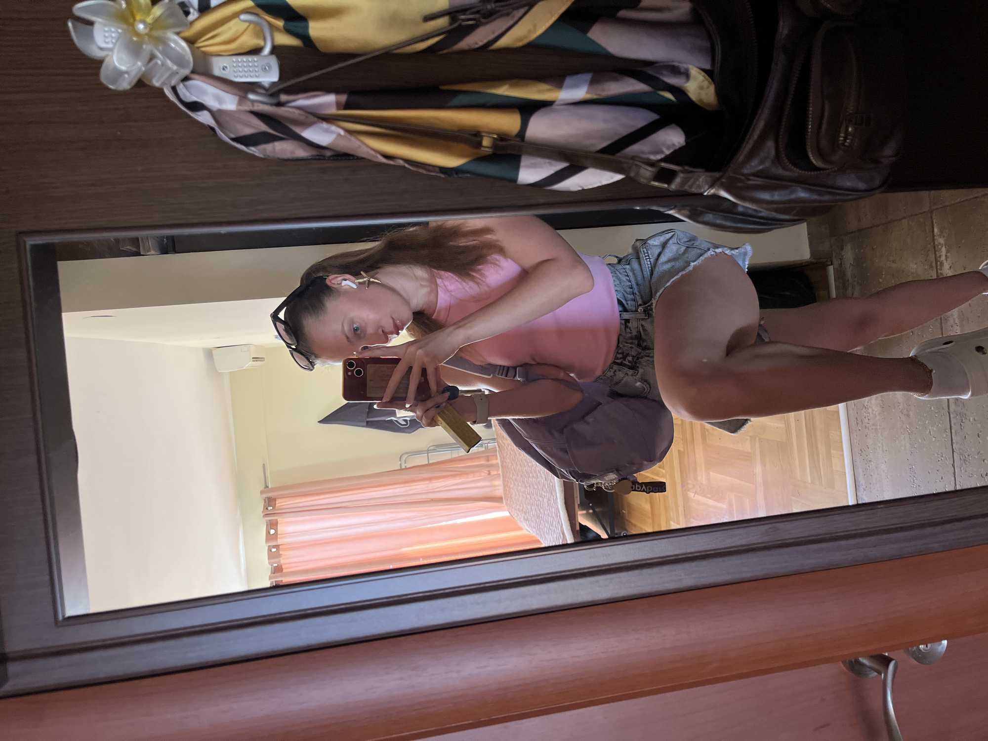 The image depicts a woman taking a selfie in a mirror. The woman is wearing a pink tank top and denim shorts. She is holding a phone in her left hand and has a pair of sunglasses perched on her head. She also appears to be wearing wireless earbuds. The reflection in the mirror shows a bedroom setting with light pink curtains and a bed, suggesting the room is interior. Above the mirror, items including a handbag, a patterned scarf or shawl, and a decorative flower are placed on what appears to be a dresser or a hanging area. It is not possible to confirm that this image was taken in Bečići, Montenegro.