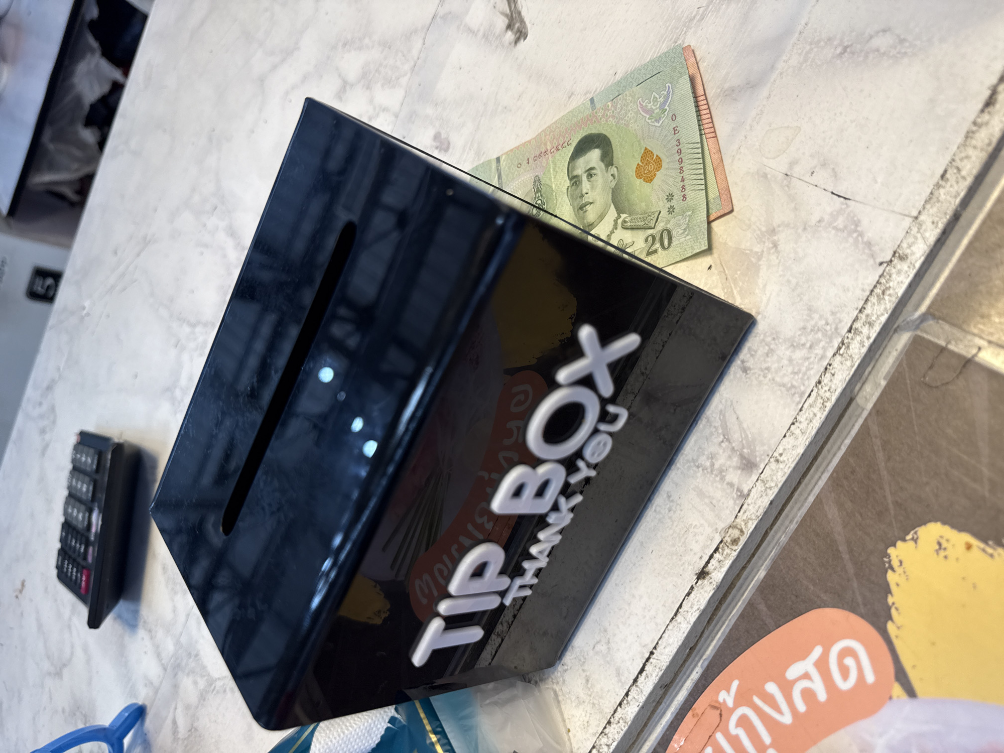 A black, rectangular tip box with white text that reads "TIP BOX THANK YOU" sits on a speckled, light-colored countertop. A Thai 20-baht note is placed next to the tip box. In the background, a black calculator is visible on the countertop. The scene appears to be indoors, possibly in a shop or restaurant in Baan Bo Phut, Thailand.