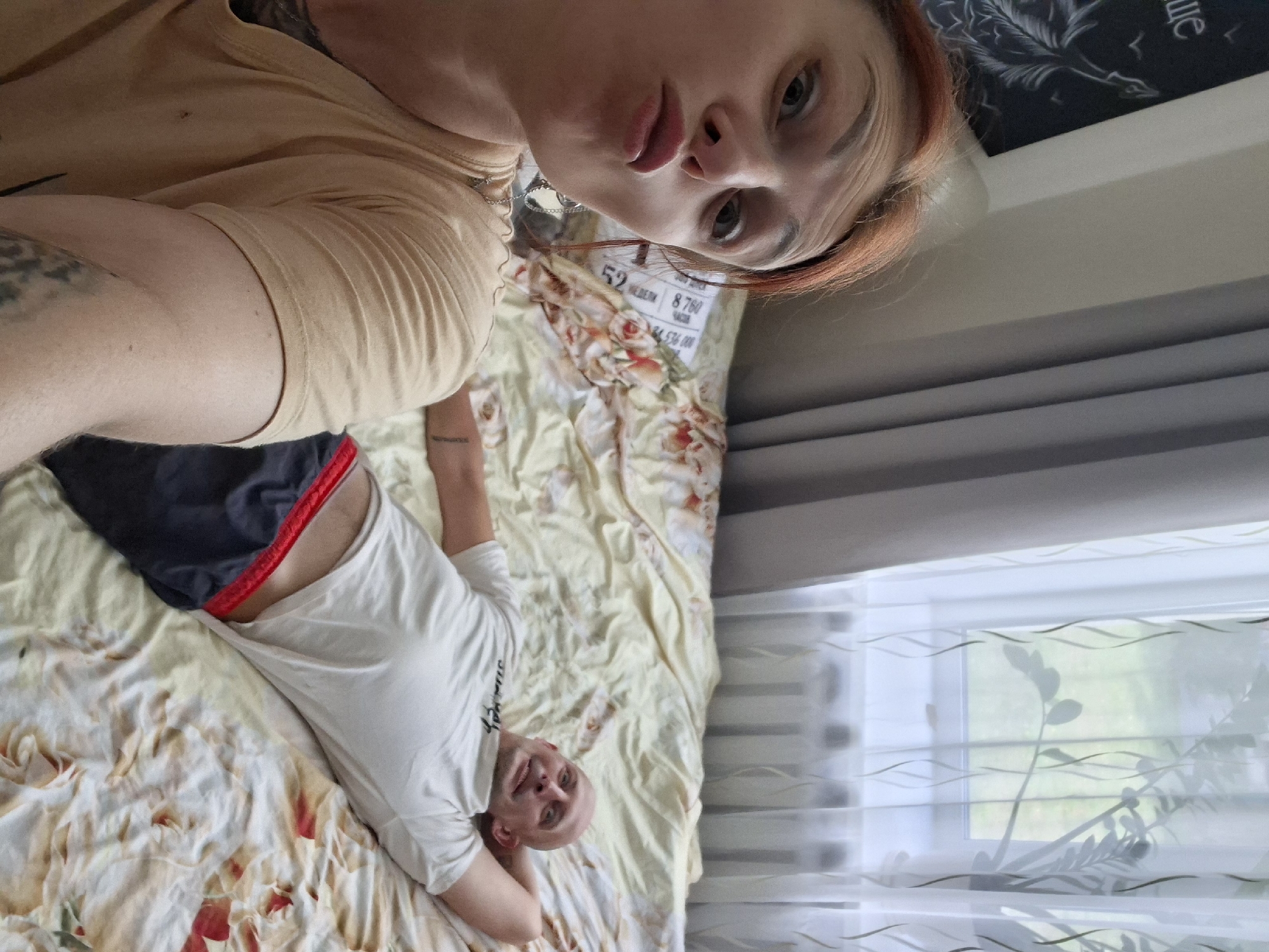 A selfie is taken by a woman with auburn hair, who is leaning towards the camera with her lips pursed. She is wearing a tan t-shirt. In the background, a bald man lies on a bed with a floral comforter. He is wearing a white t-shirt and dark shorts with a red waistband, and has his hand behind his head. The room is lit by natural light coming from a window with sheer curtains. A drawing with a bird and text is visible on the wall to the left of the woman. The scene appears to be indoors, likely a bedroom, and the presence of the drawn picture on the wall suggests it could be in an apartment or house. The overall atmosphere is casual and relaxed. The provided context indicates the location is Myadzyel, Belarus.