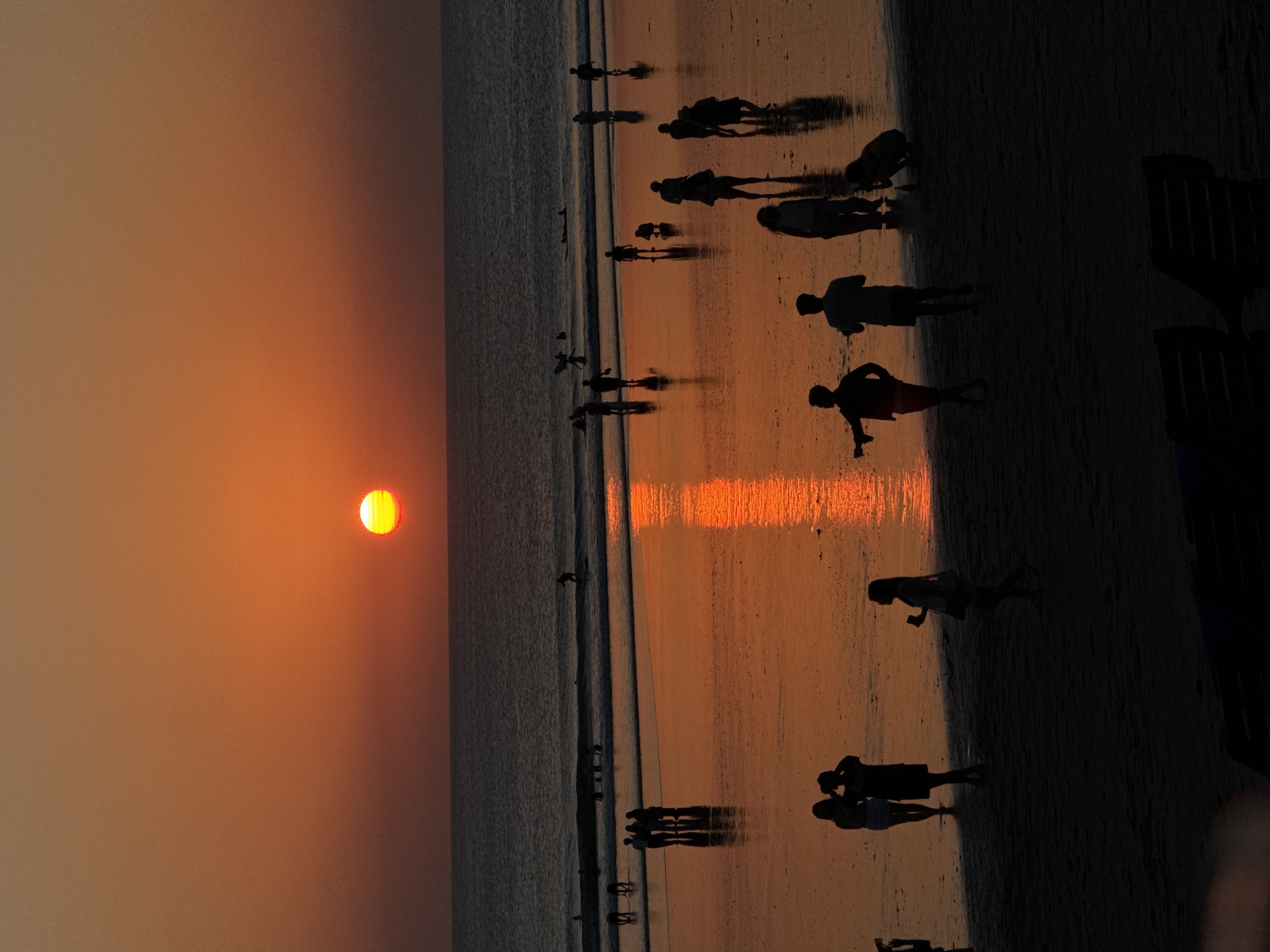 The image depicts a sunset on Kuta Beach, Bali, Indonesia. Silhouetted figures of people are visible on the wet sand and in the shallow water, some walking, some standing, and a few are in the ocean, possibly surfing. The sun, a bright orange orb, is low on the horizon, casting a warm glow across the sky and its reflection on the wet sand. The ocean stretches out to the horizon, with gentle waves. The overall scene conveys a serene and peaceful atmosphere characteristic of a beach sunset.
