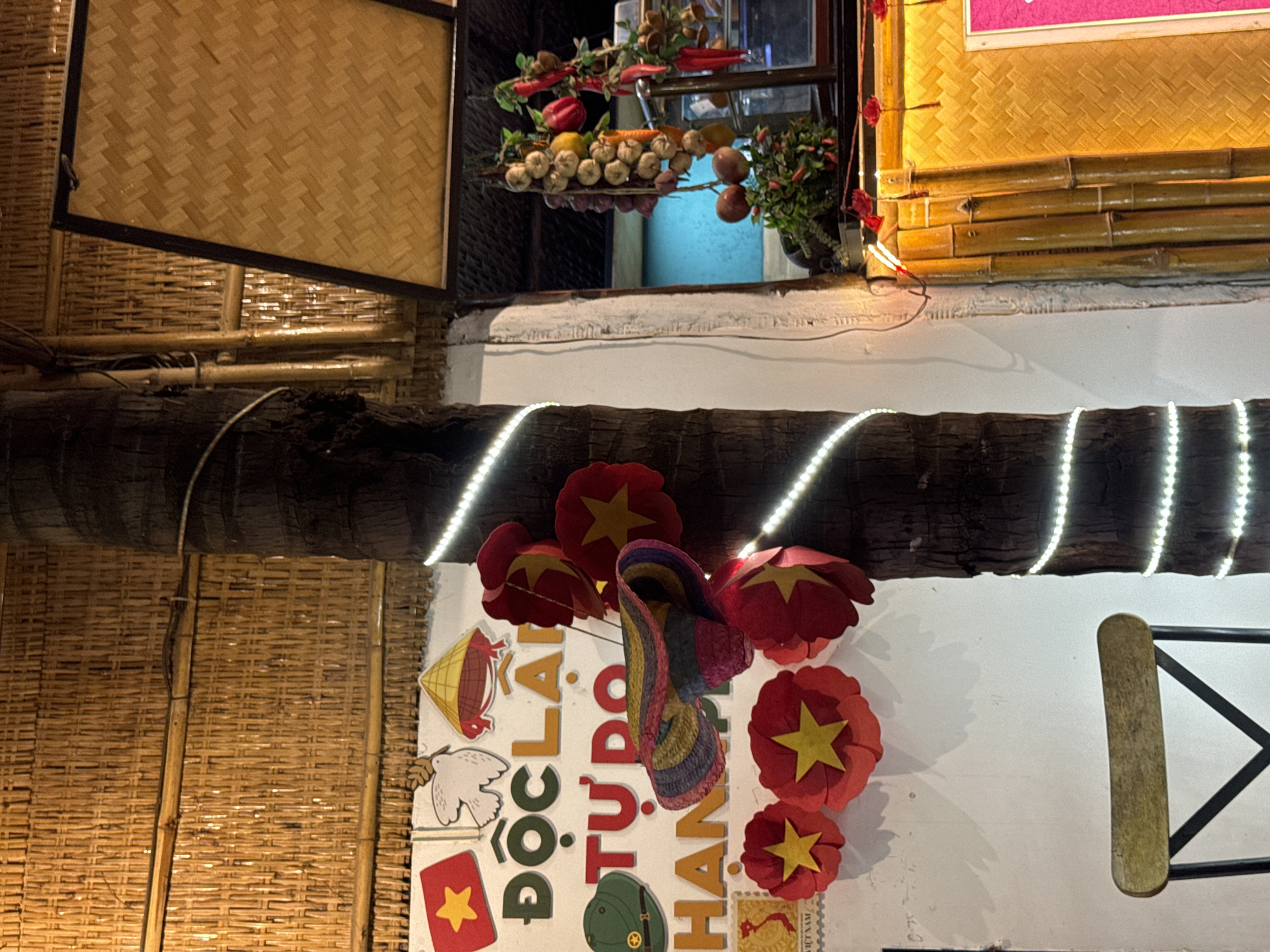 The image shows an outdoor seating area at a restaurant in Phu Thuy Ward, Vietnam. The decor features a prominent palm tree wrapped in white string lights, and a wall adorned with Vietnamese national symbols. Among these symbols are a red flag with a yellow star, a white dove, a conical hat, and text reading "ĐỘC LẬP TỰ DO HẠNH PHÚC" (Independence - Freedom - Happiness). Red paper flowers with yellow stars are also visible, along with a colorful woven hat and a military-style helmet. In the background, there are hanging decorations of garlic, onions, lemons, and chilies, as well as a yellow woven wall panel. A portion of a wooden chair is visible in the foreground. The lighting suggests it is evening or nighttime.