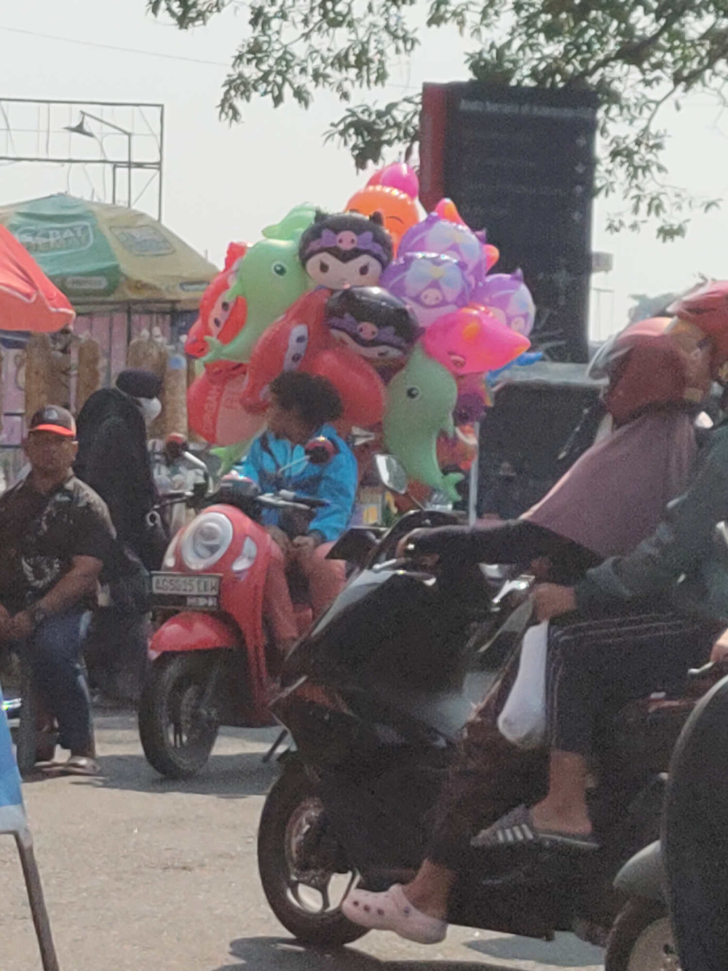 The image depicts a street scene in Ngasem, Indonesia, likely a market area. In the foreground, two motorcycles are present. The red motorcycle on the left has a person wearing a blue jacket and a helmet on it, facing towards the right. The license plate on the red motorcycle reads "AG 5915 LKW". A man wearing a black shirt and an orange cap is standing to the left of the red motorcycle, looking towards the right. Behind the man, another person in black is standing.
A prominent cluster of colorful balloons, including character-shaped ones like Hello Kitty and various animal shapes such as dolphins and dinosaurs, is attached to the red motorcycle. To the right of the red motorcycle, another motorcycle is partially visible with a person wearing a maroon helmet and a dark top.
In the background, several market stalls are visible, including a green and yellow tent with the word "SEPATT" partially visible, suggesting a shoe seller. Trees with green foliage hang over the scene, and in the far background, a large black structure, possibly a speaker or billboard, is visible against a bright sky. The ground appears to be asphalt or a similar paved surface, indicative of a road or marketplace. The overall impression is a busy, everyday scene in a commercial area.