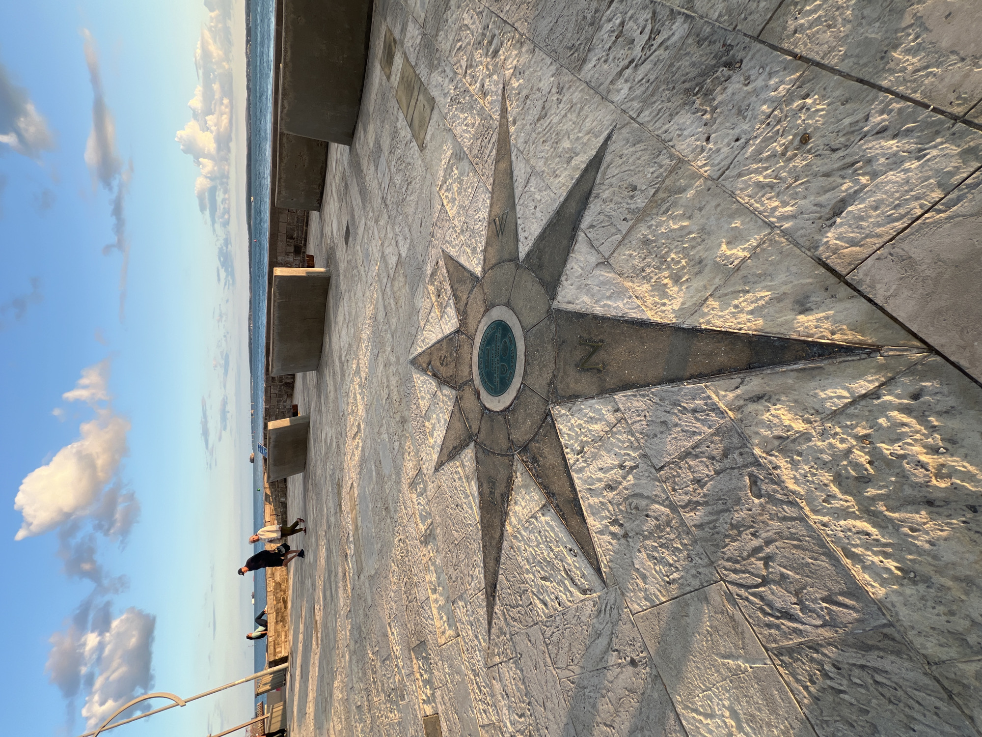 The image shows a stone-tiled promenade next to a body of water, likely the sea. A compass rose is inlaid into the paving stones, with the cardinal directions marked. To the left of the compass rose, two people are walking along the promenade. The sky is blue with scattered white clouds. The sun appears to be low in the sky, casting long shadows and illuminating the textured stone. The scene suggests a pleasant late afternoon or early morning in a coastal location. The location is identified as Portsmouth, United Kingdom. No other visible text is present in the image.