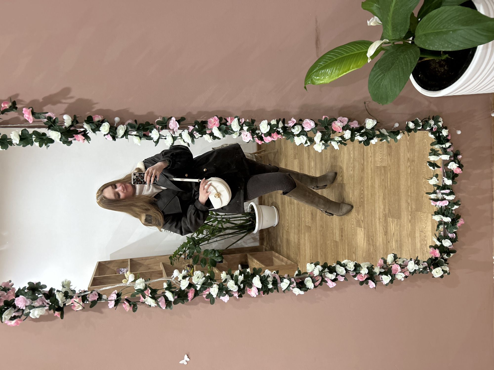 A young woman is captured in a mirror selfie, holding her smartphone up to take the photo. She is smiling slightly, her long, light brown hair falling over her shoulders. She wears a dark jacket or coat over what appears to be a light-colored top, dark leggings or pants, and knee-high, light brown or taupe boots. A white, circular handbag with a gold clasp hangs from her left arm. The smartphone has a patterned case, possibly leopard print.
The large, rectangular mirror is adorned with a decorative artificial garland of pink and white roses or blossoms intertwined with green leaves, framing its perimeter. The reflection shows a light wooden floor and a white wall. Behind the woman, inside the reflection, is a potted green plant and a multi-compartment wooden shelving unit.
Outside the mirror's reflection, the wall is a solid dusty pink or rose color. In the upper right corner, another large potted plant with broad green leaves and a small white bloom is visible. A small white butterfly decal is stuck to the pink wall below the mirror. The lighting is soft and even, suggesting an indoor setting during the day or with good artificial light. There is no visible text in the image. The scene is located in Chaikovsky, Russia, and suggests a stylish interior, possibly a boutique, cafe, or well-decorated home.