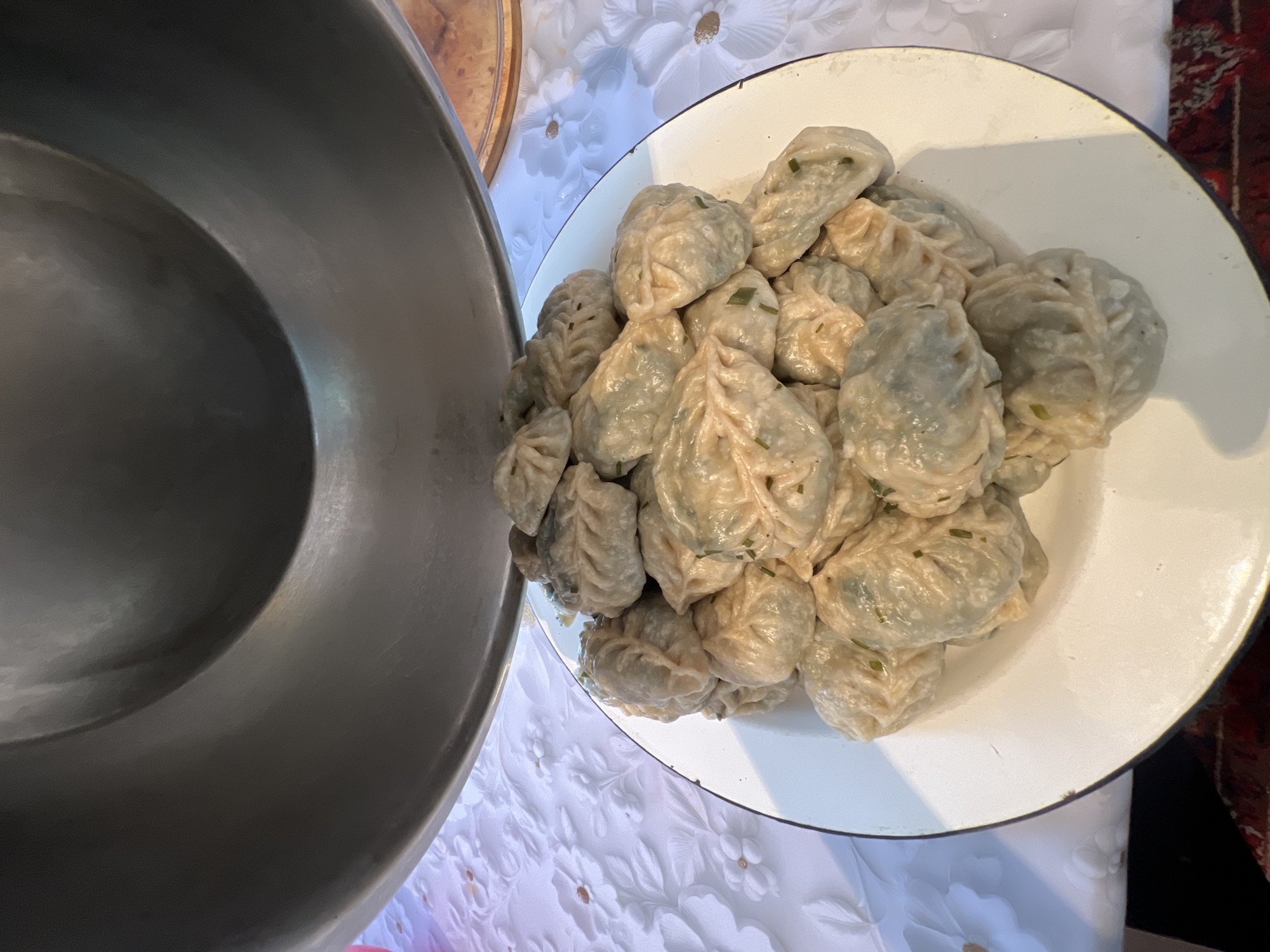 The image shows a close-up of a large white enamel plate filled with a pile of steamed dumplings. The dumplings are light-colored and have a pleated appearance, with some showing flecks of green herbs, likely chives or scallions, sprinkled on top. To the left of the plate is a large, dark-colored metal bowl, possibly a serving dish or a pot. Both the plate and the bowl are resting on a white tablecloth with a subtle floral embossed pattern. In the background, there are hints of other decorative elements, including a dark red textile with intricate patterns, suggesting a domestic setting. The lighting appears to be natural and soft, indicating it could be daytime. The scene is likely from Ak-Beshim, Kyrgyzstan, judging by the context provided. The focus is entirely on the food, suggesting the preparation or presentation of a meal. There is no visible text in the image.