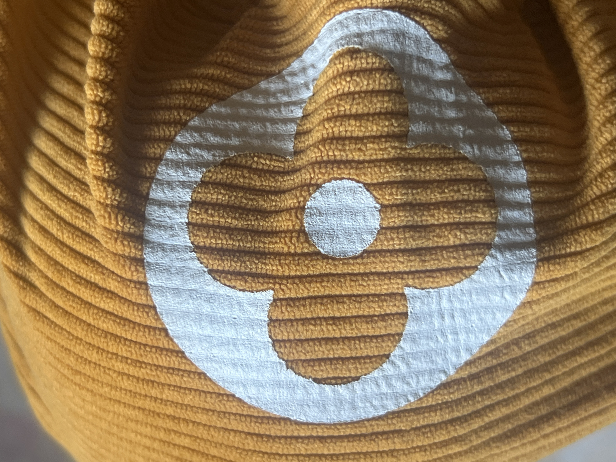 The image shows a close-up of a textured orange fabric, possibly a beanie or scarf, with a white, abstract floral or clover-like logo printed on it. The fabric has raised vertical ribs, creating a corduroy-like texture. The white logo is stylized, with a central circular element and four petal-like shapes extending outwards, all enclosed within a curved outer shape. The lighting casts shadows that highlight the texture of the fabric and the edges of the logo. No specific location in Jauro Yinu, Nigeria can be determined from this image, as it is a close-up of a textile object.