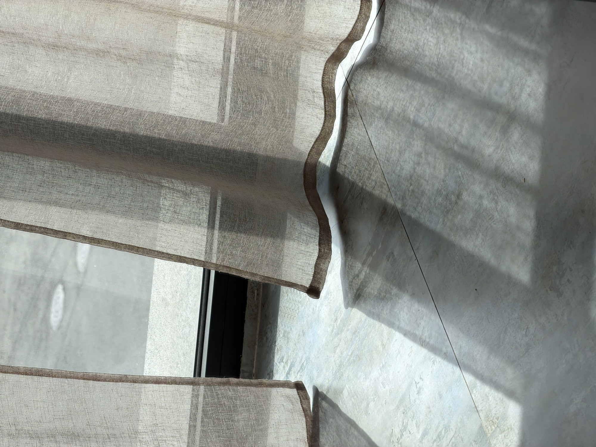 This image presents a serene and abstract indoor scene, likely within a modern building in Yerevan, Armenia. The primary focus is on translucent, light-colored fabric blinds, which allow sunlight to filter through and create intricate patterns of light and shadow.
The main subjects are the sheer, woven fabric blinds, possibly linen or a similar material, displaying a subtle texture. These blinds are partially drawn, revealing a dark window frame and a glimpse of what appears to be a light-colored outdoor or adjacent surface through the glass. The right edge of the blinds features a distinct, wavy border in a slightly darker brown tone, adding a decorative element and creating a dynamic line against the wall.
Strong, angular shadows are cast across the light gray, textured wall and the similarly colored tiled floor. These shadows are sharp and well-defined, indicating direct sunlight and suggesting a clear, sunny day. The angle of the shadows implies either morning or late afternoon, as the sun is not directly overhead. The wall exhibits a subtle, uneven texture, while the floor is composed of large, light-toned tiles with visible grout lines.
The scene is devoid of people or activity, conveying a sense of calm and quiet observation of natural light interacting with architectural elements. The interplay of light, shadow, and texture creates a minimalist aesthetic. There is no visible text in the image.
