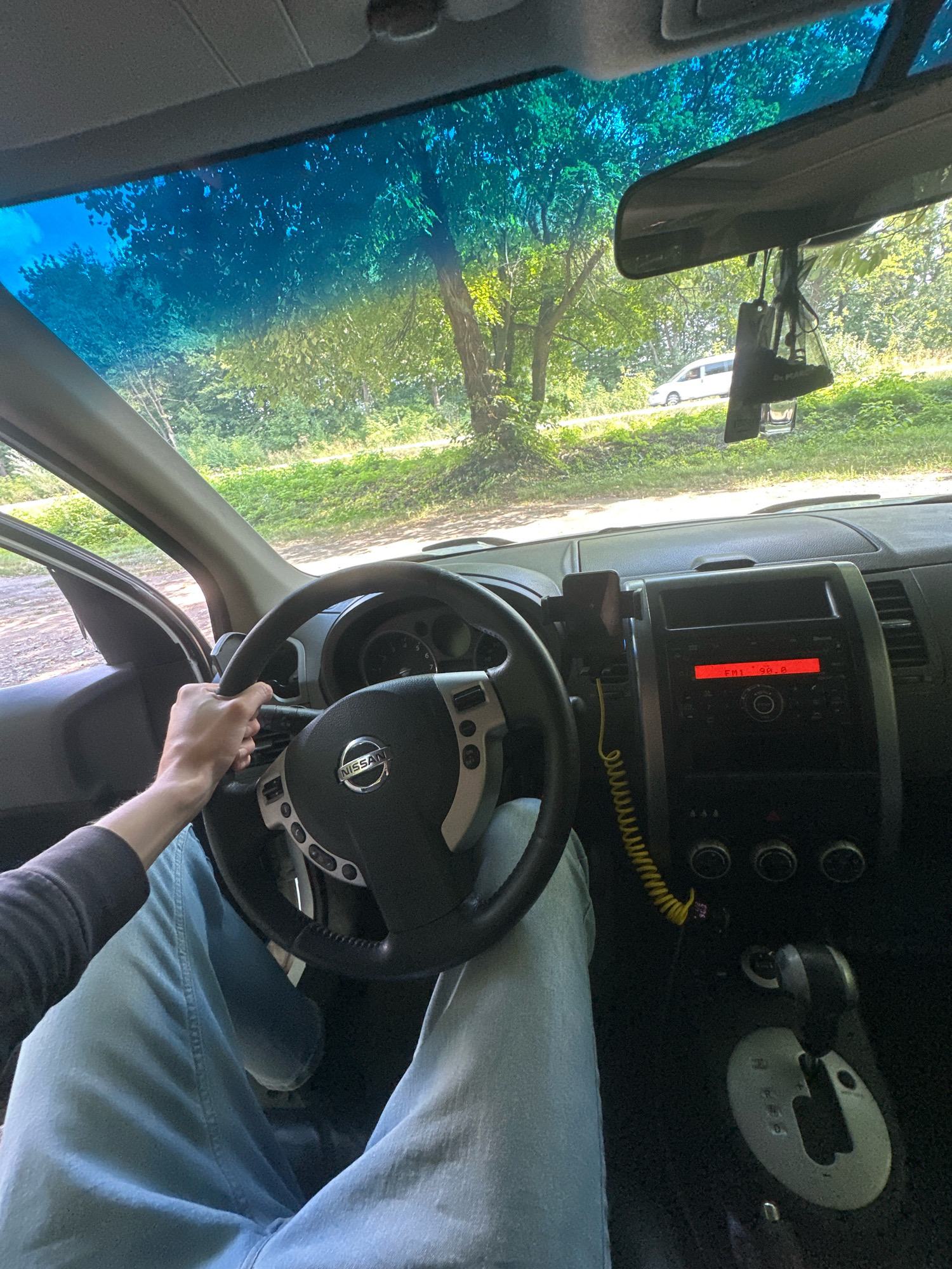 The image is a first-person perspective from inside a Nissan car, with the driver's hands on the steering wheel. The driver is wearing light blue jeans and a dark long-sleeved shirt. The dashboard shows a radio displaying "FM1 98.8" and a gear shift. Outside the car, through the windshield and side window, a tree-lined road is visible with green foliage. A white vehicle is parked or driving on the road in the distance. A rear-view mirror with a car freshener hanging from it is also visible. The scene appears to be in a rural or suburban area. The location is identified as Shatava, Ukraine.