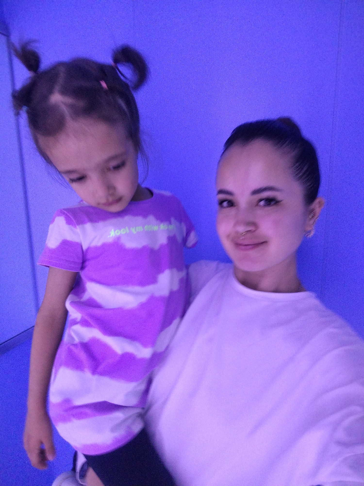 The image shows two females, an adult and a child, posing for a selfie against a solid purple background. The adult, on the right, is looking directly at the camera with a slight smile. She has dark hair pulled back in a bun and is wearing a white shirt. The child, on the left, is being held by the adult and is looking down. She has dark hair styled in two pigtails and is wearing a purple and white horizontally striped t-shirt. The environment appears to be indoors, possibly a studio or a room with purple-colored walls and floor. There is no visible indication of the city of Chaikovsky, Russia.