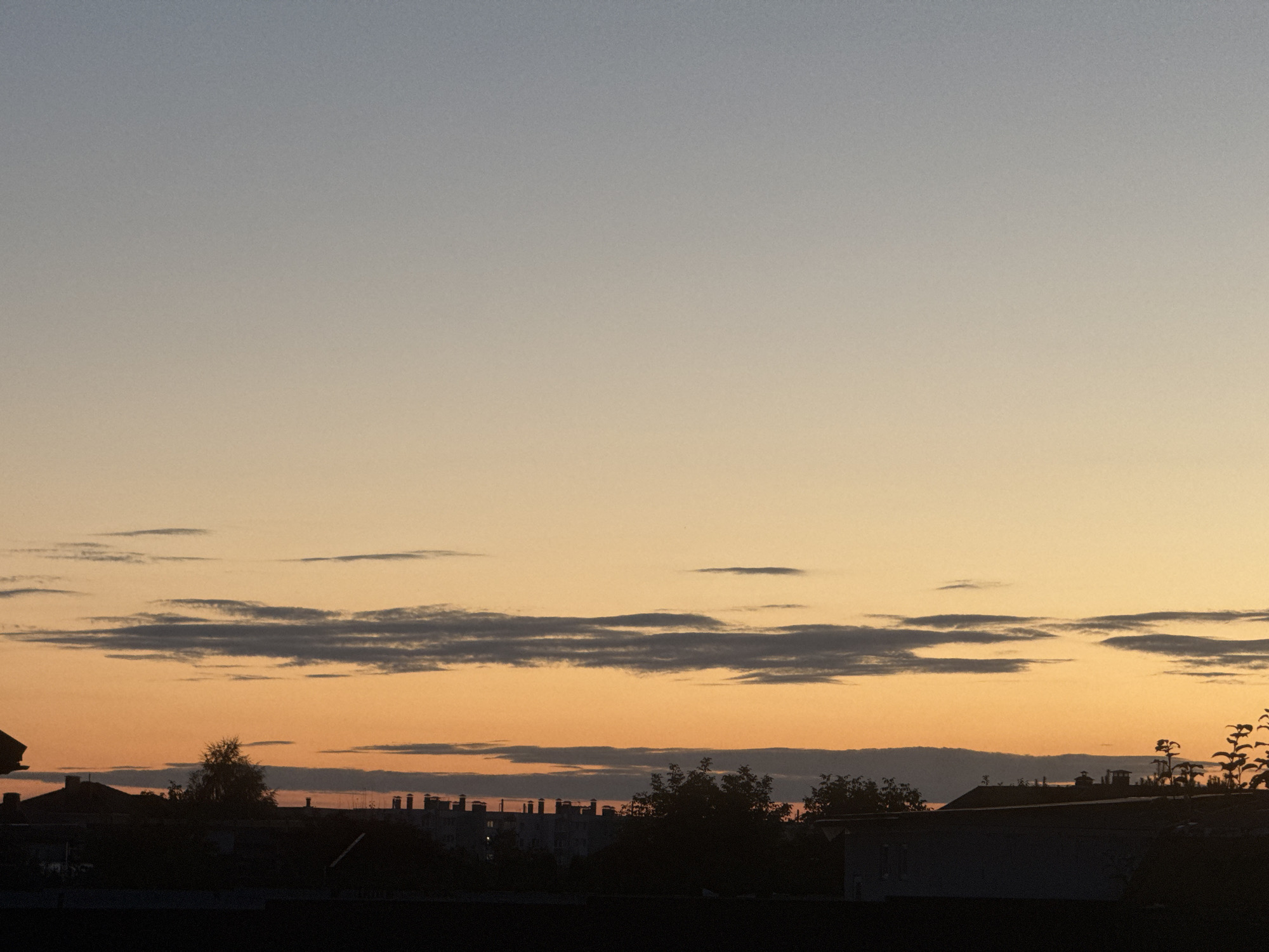 The image shows a silhouetted cityscape against a gradient sky transitioning from pale blue at the top to a warm orange near the horizon. Wispy, dark clouds are scattered across the mid-to-upper portion of the sky. In the foreground, the dark silhouettes of buildings, trees, and rooftops are visible, suggesting an urban or suburban environment. The scene appears to be captured during either sunrise or sunset. The specific location within Бараўлянскі сельскі Савет, Belarus is not discernible from the visual information alone.