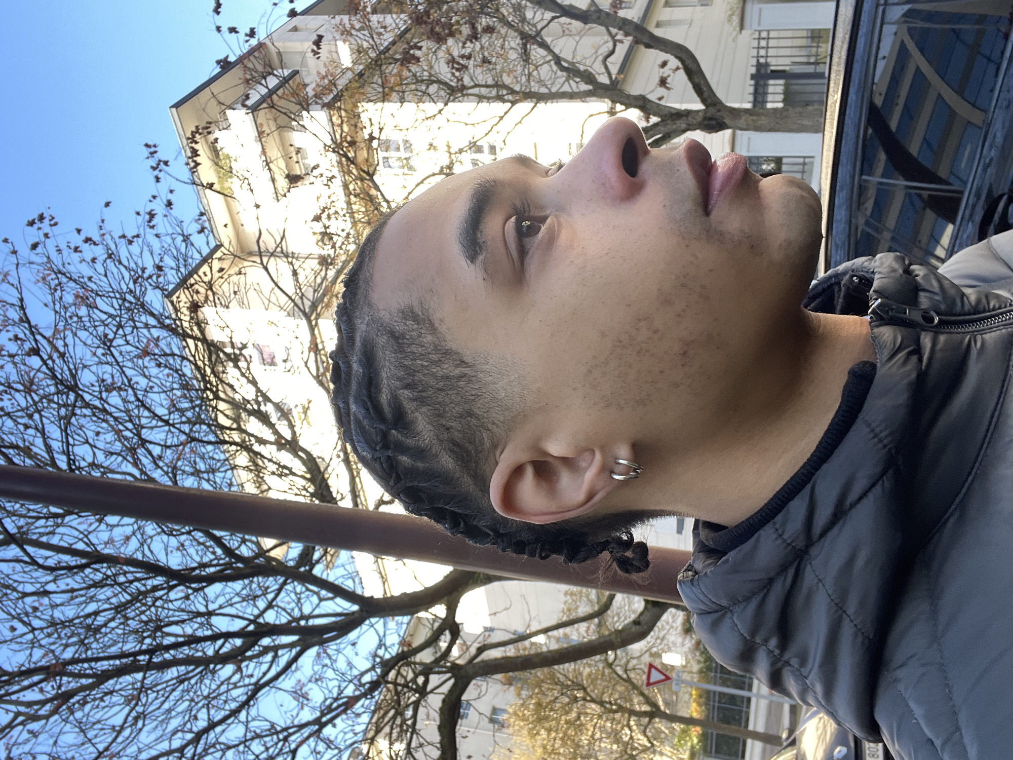 An individual, identified as a young male, is depicted in profile from the chest up, facing right, in Montigny-le-Bretonneux, France. He possesses dark skin, a short fade haircut on the sides, and dark cornrows styled on top. His left earlobe features two silver hoop earrings. Facial details include brown eyes, a defined nose, full lips, and a light stubble on the chin and jawline, with scattered minor epidermal blemishes on his cheek. He is attired in a dark, possibly grey or black, quilted puffer jacket over a dark, high-collared inner garment.
The background presents a clear, bright blue sky. Numerous deciduous trees, largely bare with sparse reddish-brown leaves, are visible, suggesting an autumn or winter setting. A tall, dark brown pole bisects the upper left portion of the frame. Behind the trees, multiple multi-story residential buildings with light-colored facades, flat or gently sloped grey roofs, and visible balconies are illuminated by sunlight. To the left, a portion of a red triangular 'yield' traffic sign is visible. In the lower right, a dark-colored vehicle reflects the surrounding environment. To the lower left, the front section of a lighter-colored vehicle is partially visible, displaying the initial characters "80" of a license plate. The overall scene indicates an urban outdoor environment under daylight conditions.
