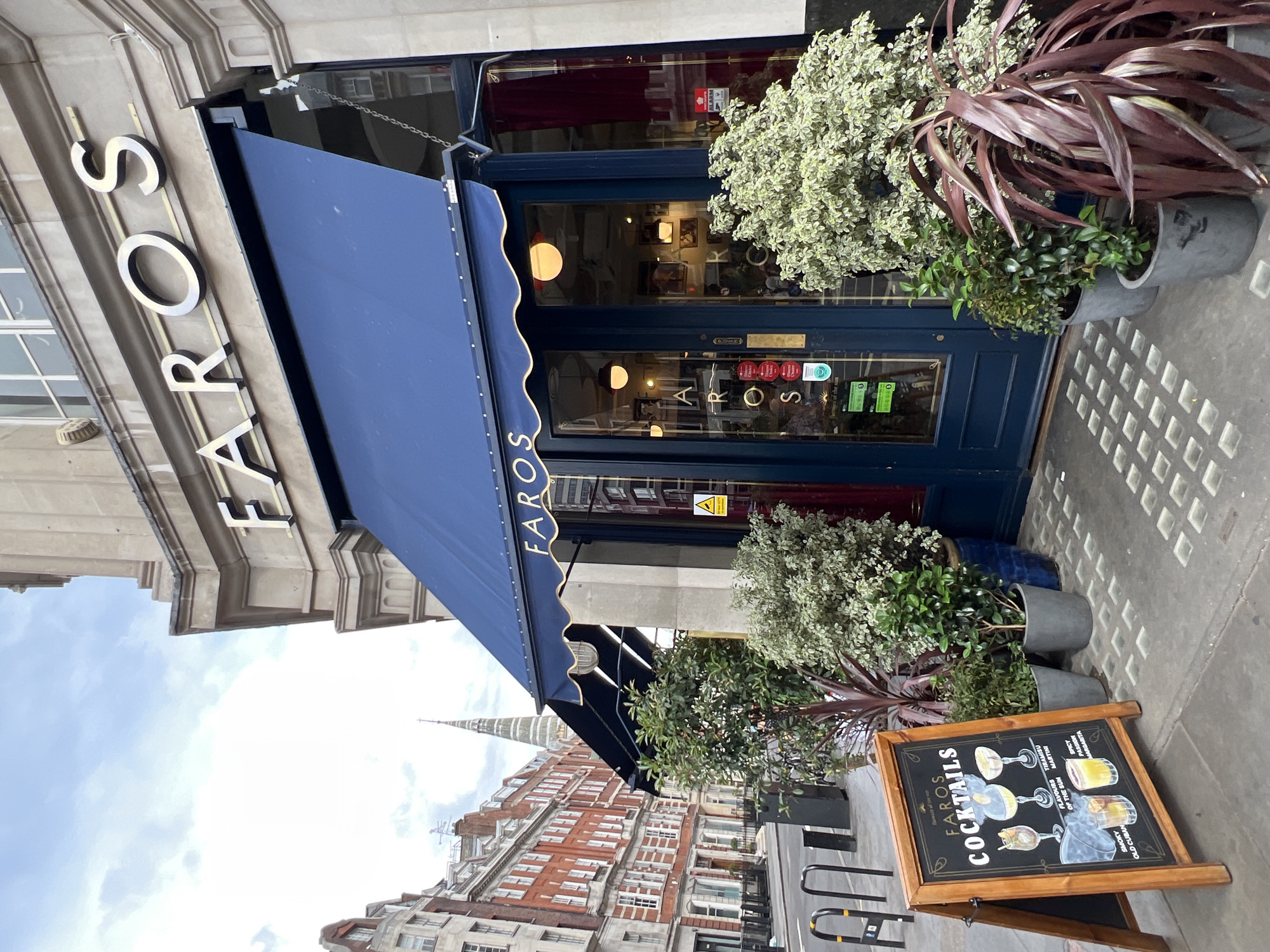 The image shows the exterior of a restaurant named FAROS, with a prominent blue awning over the entrance. The restaurant's name is also displayed in large silver letters above the awning. The entrance consists of double blue doors with large glass panes, revealing the interior of the establishment. In front of the entrance, there are several potted plants, adding greenery to the scene. To the left of the entrance, a wooden A-frame sign displays an advertisement for FAROS COCKTAILS, with illustrations of various drinks. In the background, a street scene unfolds, featuring multi-story buildings with traditional architecture, including red brick facades and white-trimmed windows, and a spire of a church or tower piercing the sky. The overall environment suggests an urban setting, likely a city street with historical buildings. The sky is overcast.