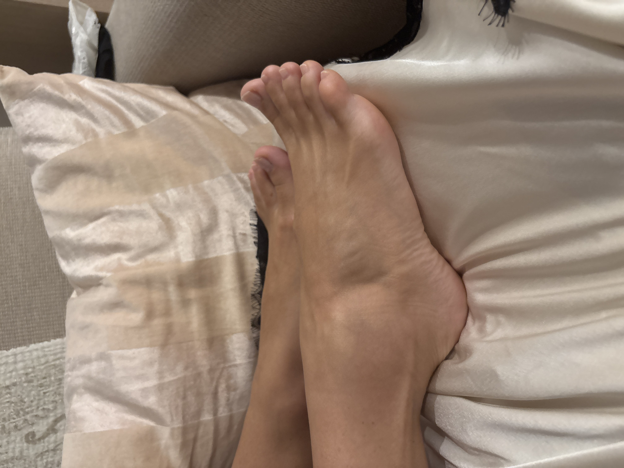 The image shows a close-up view of a person's bare feet resting on a cream-colored fabric. The fabric appears to be satin or silk and is draped over a piece of furniture that looks like a sofa or a bed. The feet are positioned with the toes pointing towards the right side of the frame. The skin tone of the feet suggests they belong to a person with fair to medium complexion. The lighting in the image is soft, casting subtle shadows that highlight the contours of the feet and the folds of the fabric. There are no other objects or people visible in the frame. The background shows a textured pillow and the armrest of the furniture, suggesting an indoor setting, possibly a living room or bedroom. There is no indication of the location being Rome, Italy.