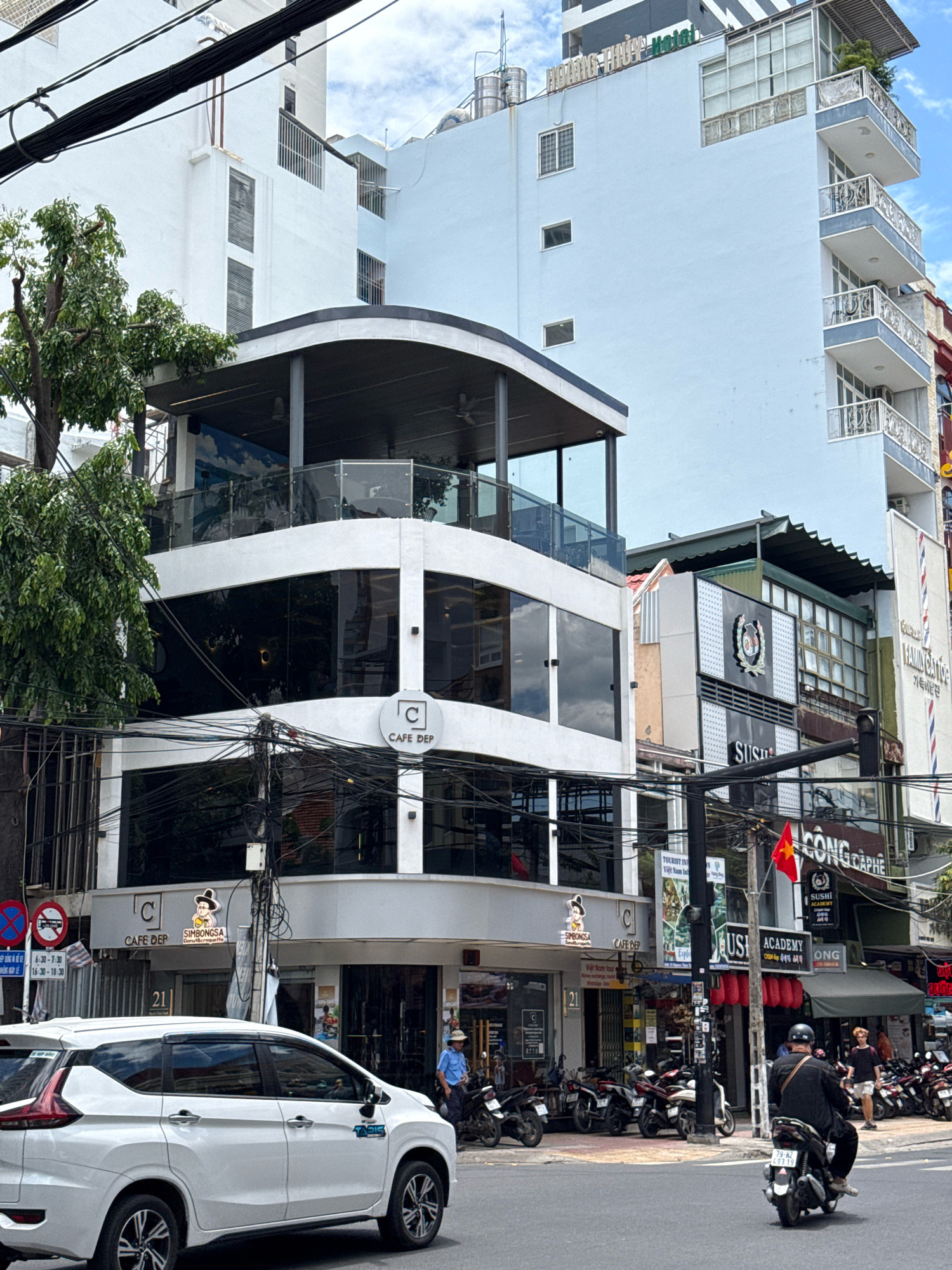 A white SUV is parked on the right side of the street in Nha Trang, Vietnam. The car is facing left and is partially obscured by a tree on the left. In the background, there is a multi-story building with balconies and windows. A street sign with Vietnamese writing is visible on the left side of the image. The sky is cloudy.