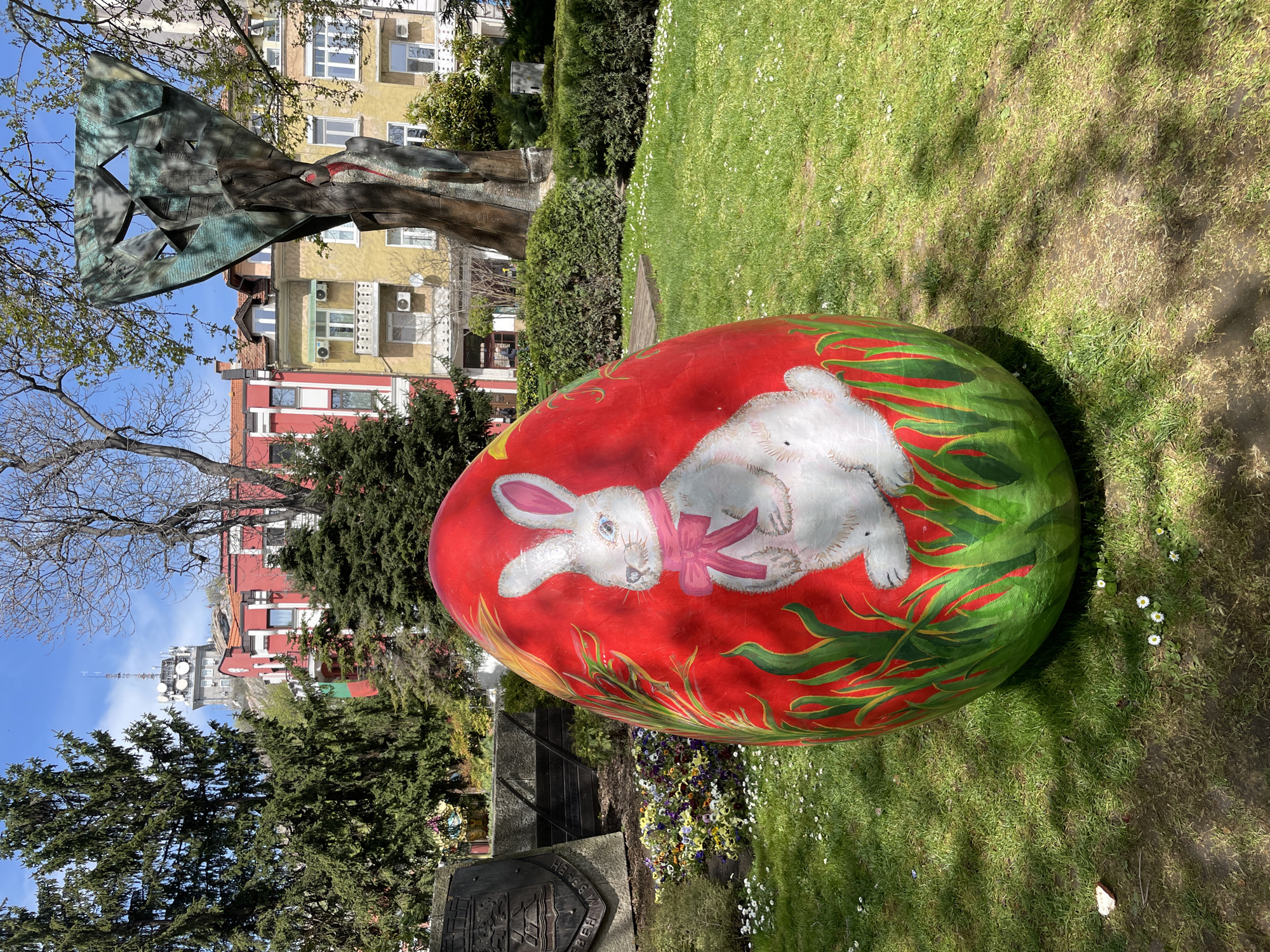 A vibrant, large, artificially-colored Easter egg is prominently displayed in a park setting, lying on its side on a patch of green grass. The egg is painted with a bright red background, featuring a white Easter bunny adorned with a pink bow, nestled among green foliage designs. Small white flowers dot the surrounding grass, indicating springtime.
In the background, a sunny day with a clear blue sky illuminates the scene. To the left, a large, abstract, greenish-bronze sculpture or monument stands amidst trees. Beyond the immediate park area, several multi-story buildings are visible; one is yellow/beige with multiple windows and air conditioning units, and another is painted red. Further in the distance, a distinctive communication tower, equipped with multiple antennae, rises above the cityscape, possibly situated on a higher elevation.
No people are visible in the image. The presence of the decorated egg strongly suggests an Easter celebration or public display, creating a festive atmosphere. Shadows cast on the grass indicate bright sunlight, placing the time of day likely between late morning and afternoon. At the base of the image, near the large egg, a stone pedestal features a crest or coat of arms, with partially legible Cyrillic text below it, including the letters "ИВ". Given the context of Plovdiv, Bulgaria, this might be a local emblem or dedication plaque.
