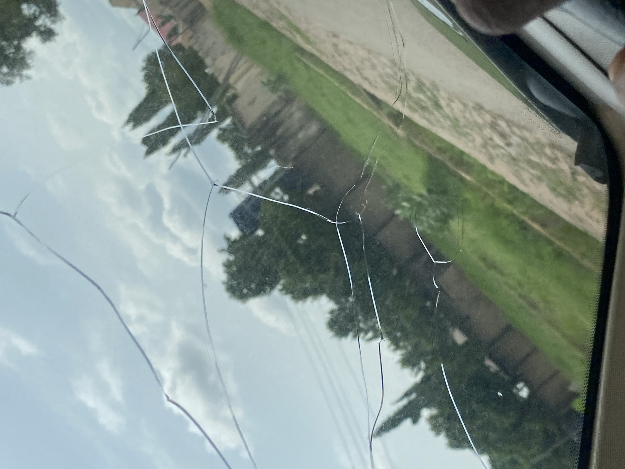 The image shows a cracked windshield of a vehicle, with the sky and trees visible through it. The scene appears to be an outdoor setting in Howayi, Nigeria. There are no people or specific actions depicted, only the damaged windshield and the environment reflected or seen through it. The cracks in the windshield are prominent and are the main focus of the image.