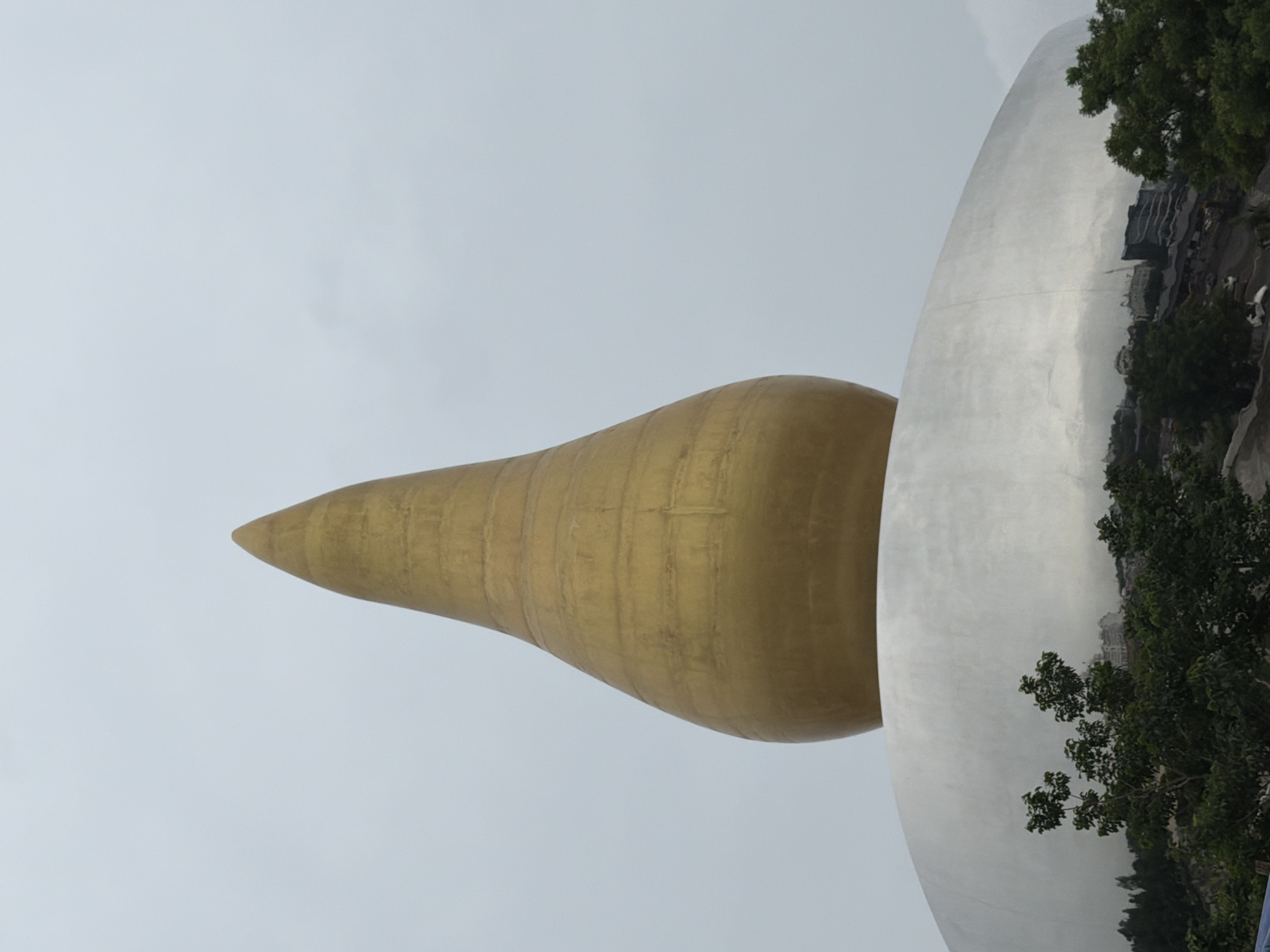 The image captures a close-up view of a large, golden, teardrop-shaped sculpture perched atop a broad, silver, dome-like structure. The sculpture is textured with horizontal bands and tapers to a pointed apex, set against a pale, overcast sky. The silver base exhibits reflections of surrounding trees and buildings, hinting at an urban environment. In the foreground, lush green trees partially obscure the base of the structure. The overall impression is one of modern architectural design, possibly a monument or a building in Hyderabad, India, given the context.