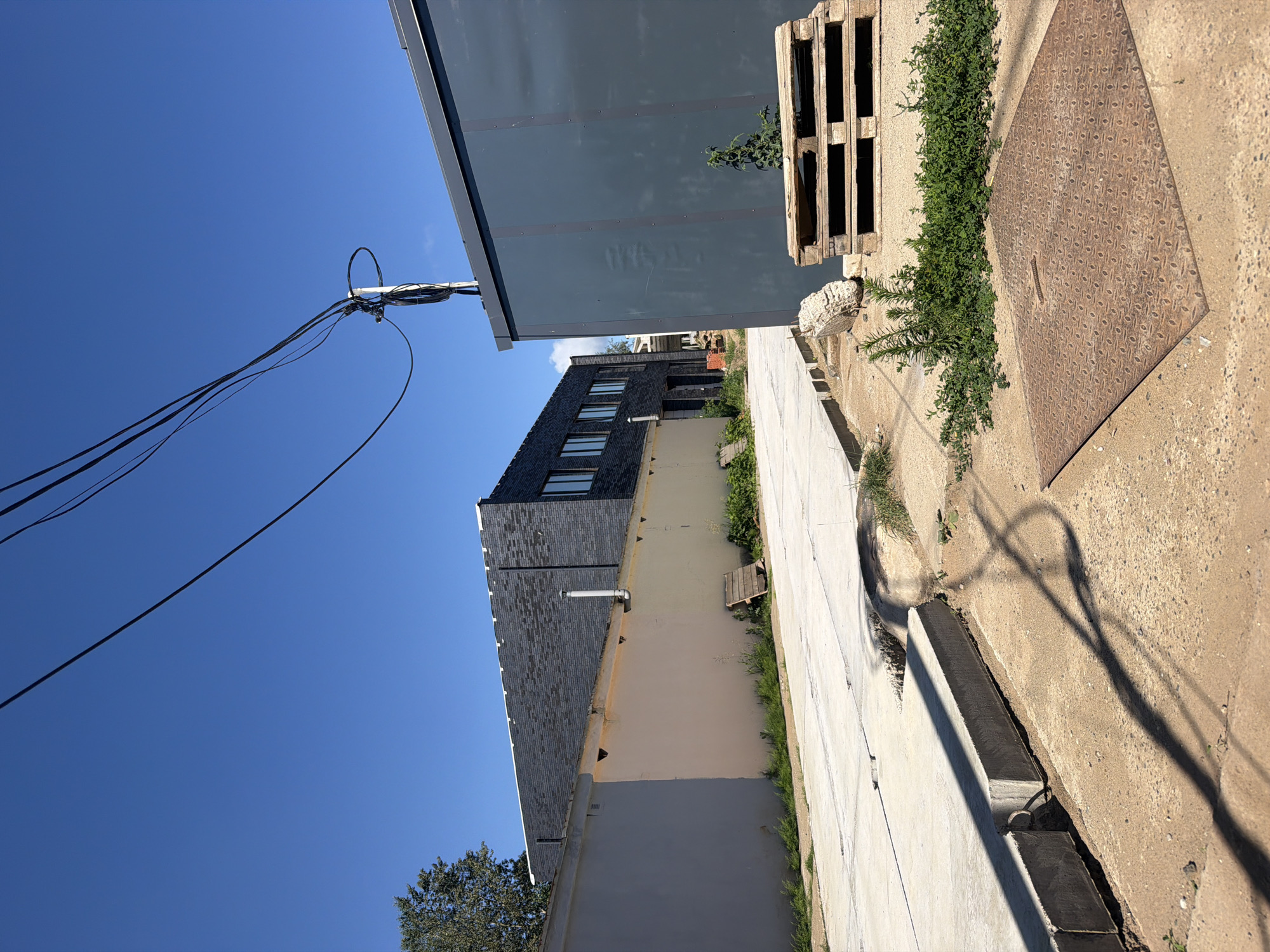 The image depicts an outdoor urban scene in Chaikovsky, Russia, under a clear blue sky. In the foreground, a textured metal manhole cover is visible on a concrete surface, with a shadow cast by overhead wires. To the left, a concrete curb runs along a paved area, with more concrete and dirt visible. Behind the curb, a light-colored wall extends into the distance. Further back, a darker, modern building with a brick facade and windows stands. To the right, a tall, dark gray structure, possibly a shipping container or modular building, is prominent. In front of this structure, a stack of wooden pallets and a large rock are present. Vegetation, including grass and shrubs, grows along the edges of the paved areas and around the structures. Overhead, black electrical wires are strung across the sky, connected to a pole. The scene appears to be an industrial or construction area.