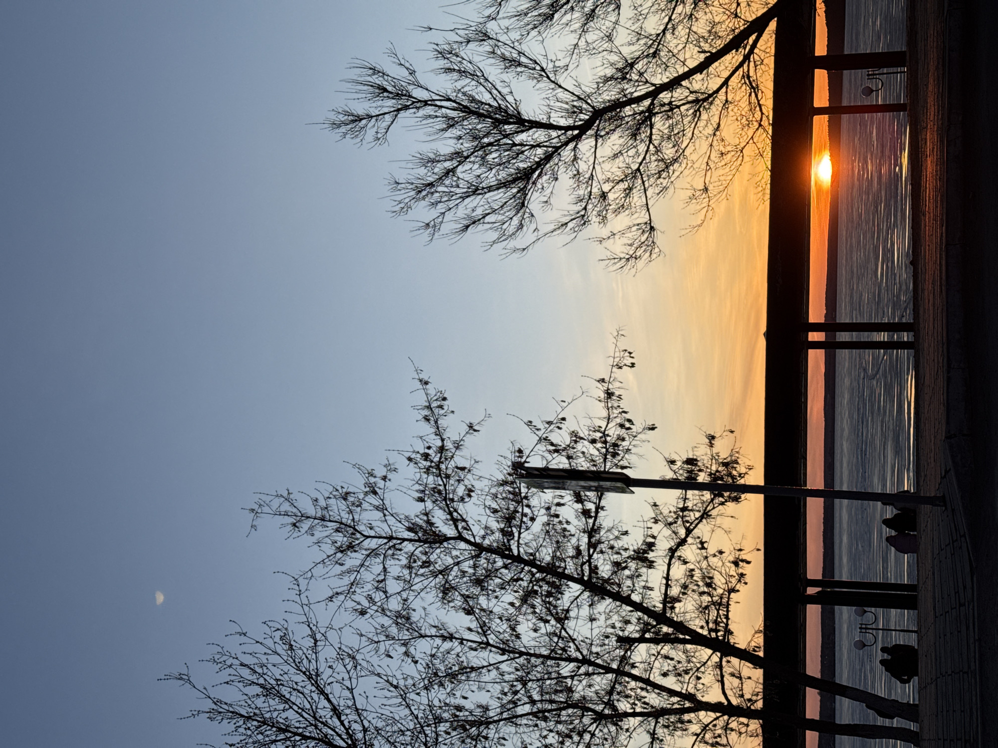 The image captures a sunset over a body of water, likely a lake or river. A portion of the skyline is colored with orange and yellow hues indicating the sunset. Silhouetted trees are visible in the foreground, framing the view of the water and the sunset. A dark structure resembling a railing or wall runs along the side of the water. There is a faint crescent moon in the upper left. Considering the request to describe the image as if it were taken in Votkinsk, Russia, and given the visible elements, it is plausible this is a view of the Votkinsk Reservoir. The dark structures could be part of a pier or boardwalk along the reservoir.