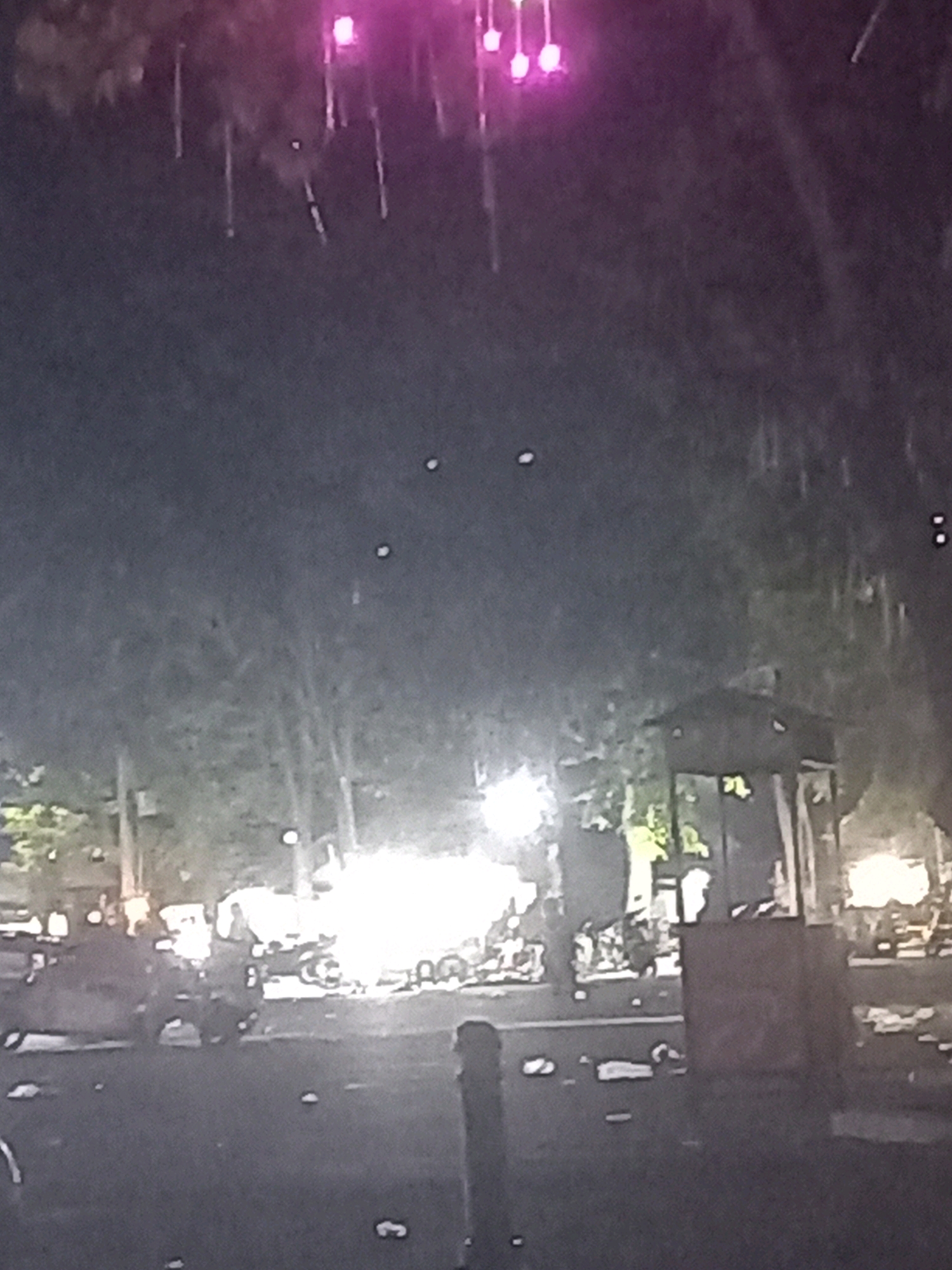 The image depicts a dark, outdoor scene, likely at night, with several elements that suggest a public gathering or event. In the upper portion of the image, there are bright pink lights, possibly from stage lighting or decorations, with some vertical elements that could be rigging or hanging fixtures. The background consists of blurred trees and foliage.
In the midground and foreground, there are hints of human activity and structures. Several bright, white lights are visible, illuminating parts of the scene. There appear to be multiple figures, some standing and others possibly seated or moving. Some of the figures are silhouetted against the lights, making detailed identification difficult.
There are also several objects scattered on the ground, some of which resemble discarded trash or debris. A prominent structure in the right midground resembles a booth or small building with a weathered appearance. Vehicles, possibly motorcycles or scooters, are also visible, some appearing to be parked or being ridden.
The ground surface is dark and appears to be paved, consistent with a street or plaza. The overall impression is of a somewhat chaotic or lively outdoor event, with a mixture of lighting, people, and environmental elements.
Based on the provided context, this scene is located in Kandangjati Kulon, Indonesia.