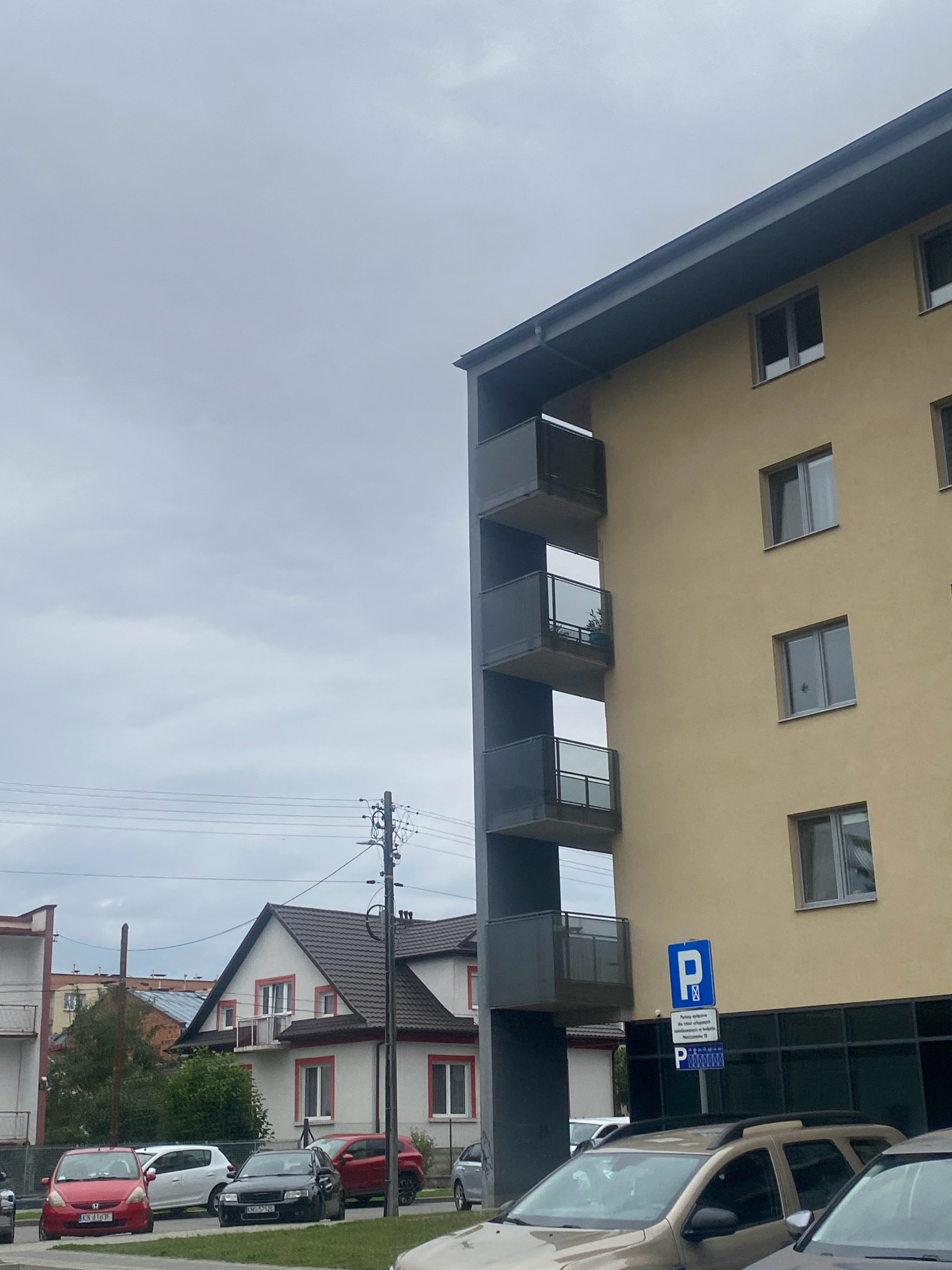 The image shows a modern apartment building with a yellow facade and dark grey balconies in the foreground, located in Nowy Sącz, Poland. To the left of the apartment building, a traditional house with a brown tiled roof, white walls, and red window frames is visible. Several cars are parked on the street in front of these buildings, including a red hatchback, a white hatchback, a black sedan, and a red SUV. A blue parking sign with a white "P" is posted on the side of the apartment building. The sky is overcast with grey clouds, and power lines and a utility pole are present in the background. The scene depicts a residential street in a city.