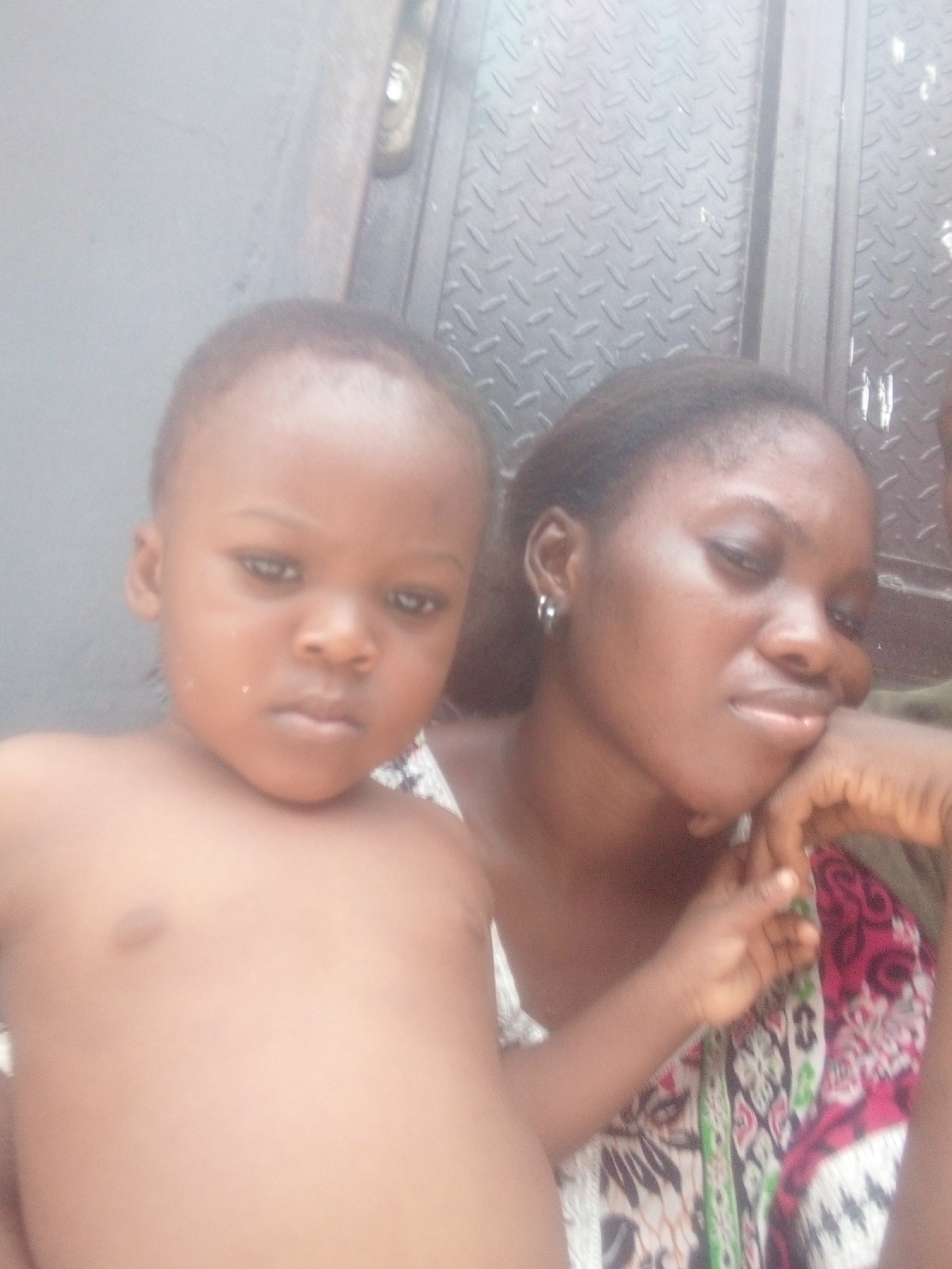 A close-up portrait shows a young child and an adult woman in an ambiguous setting. The child, positioned in the foreground and slightly to the left, has light brown skin, very short dark hair, and dark eyes, looking directly at the camera with a neutral to slightly solemn expression. The child is bare-chested.
To the right and slightly behind the child is a woman with darker skin and dark hair pulled back. She wears a vibrant patterned garment featuring pink, white, green, and black designs. A silver hoop earring is visible in her left ear. Her head is tilted slightly downwards and to the right, and her right hand rests near her chin, suggesting a moment of contemplation or relaxation, accompanied by a faint, gentle smile.
The background consists of two distinct elements. To the left, there is a smooth, light grey surface, possibly a wall or door. To the right, a textured metallic surface, resembling diamond plate, is visible, separated by a vertical seam. A small, blurry golden or brass-colored object, possibly a handle or latch, can be glimpsed at the top right. The lighting is soft and diffused, indicating an indoor or sheltered outdoor environment. No visible text is present. The overall scene conveys a calm, personal moment between the two individuals.