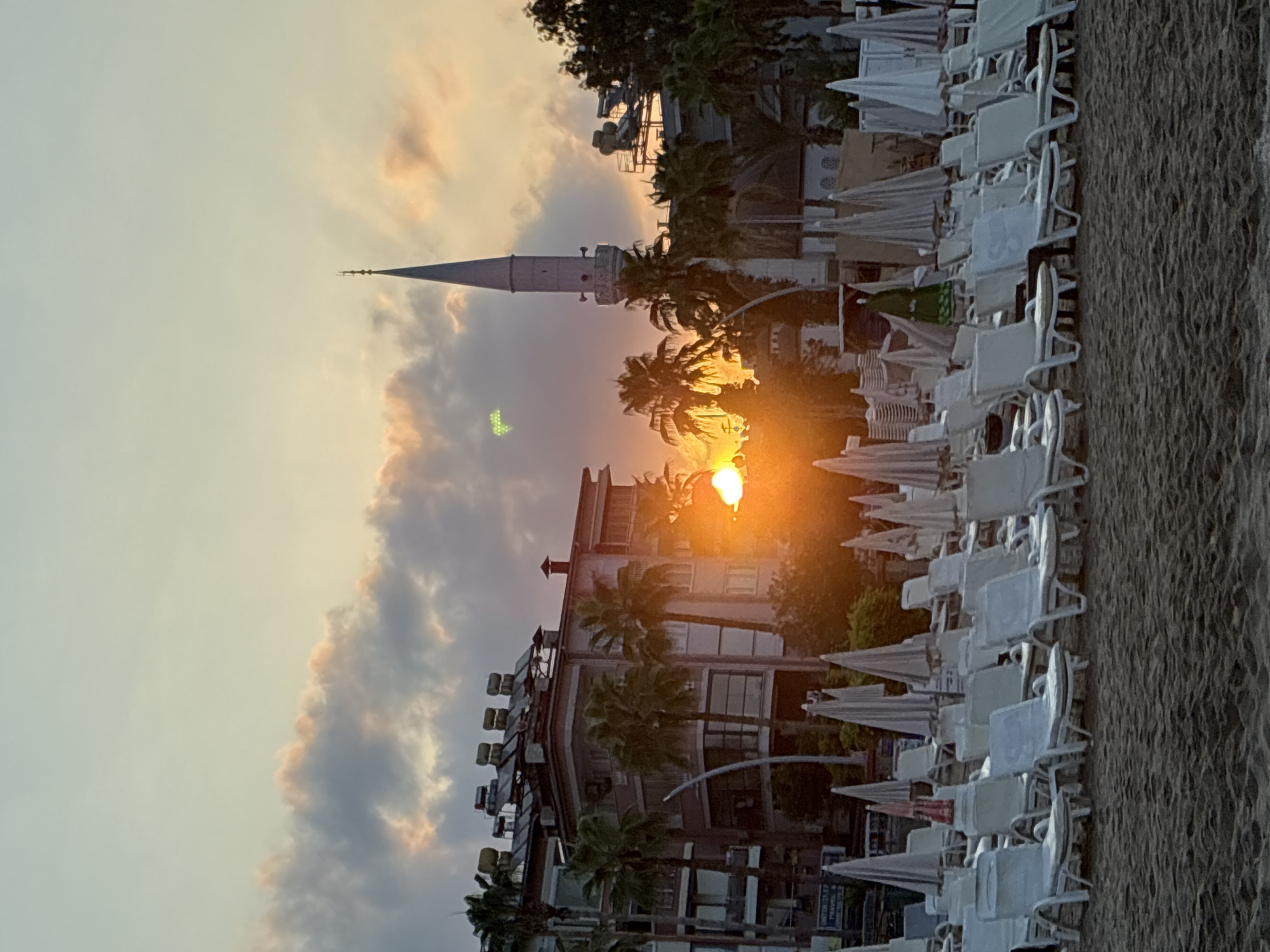 The image depicts a tranquil beach scene in Alanya, Turkey, at sunset. The sky is a soft blend of pale blue and warm orange, with scattered clouds tinged with pink and gold. The sun, a bright orange orb, is setting behind a building with solar panels on its roof and palm trees.
In the middle ground, a white minaret of a mosque rises prominently against the sky. Beside it, a tall building with balconies and palm trees in front is visible. The foreground is occupied by rows of empty white beach chairs and closed umbrellas, arranged neatly on the sandy beach. The calm sea is visible in the far background, with gentle waves lapping at the shore. The overall atmosphere is serene and peaceful, suggesting the end of a pleasant day in this coastal city.