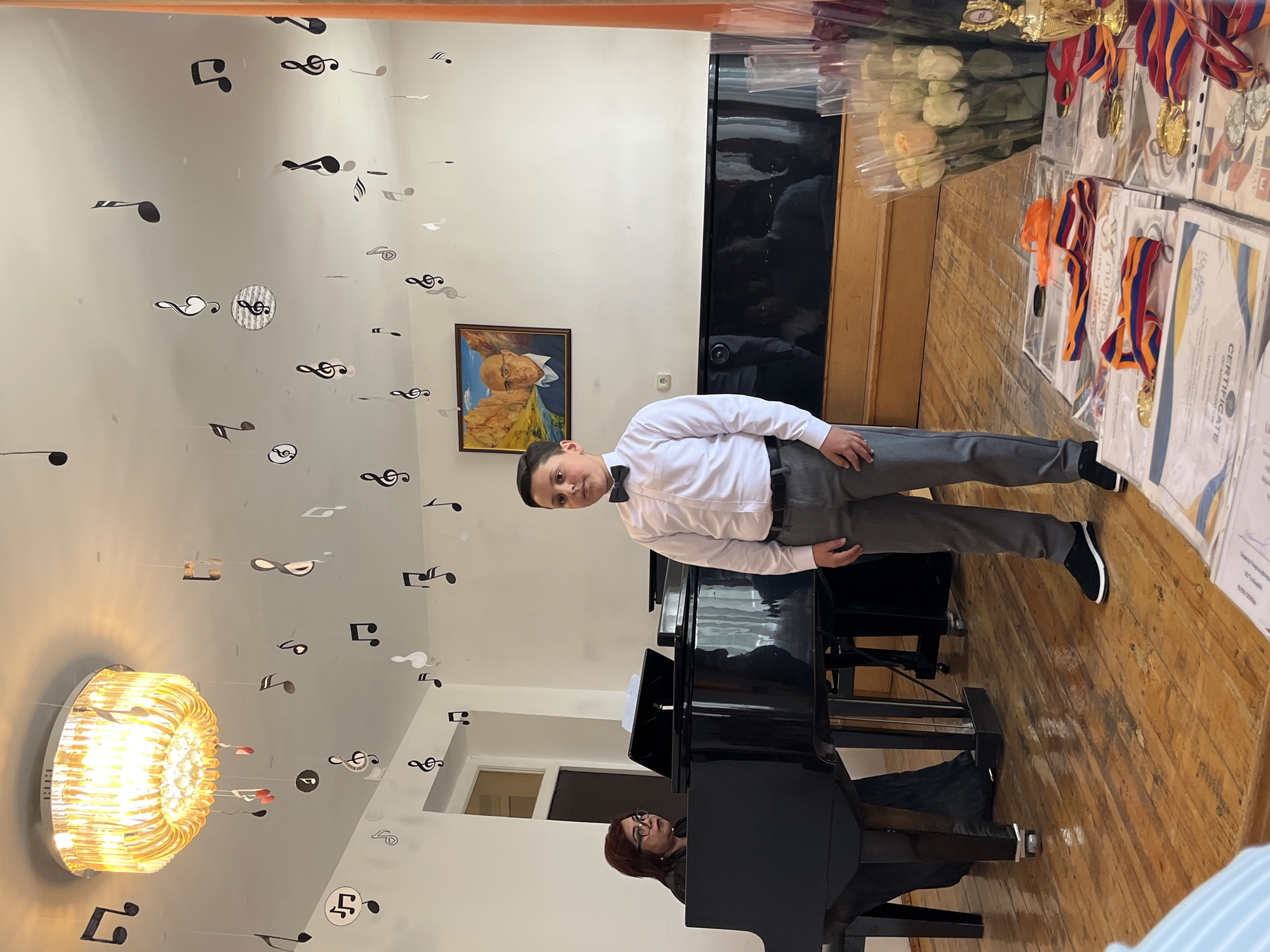 An indoor scene, likely a music recital hall, features a young boy in a white dress shirt and bow tie standing in the foreground. He is facing forward, with a neutral expression. Behind him, a woman in glasses is seated at a grand piano, looking towards the boy. The room is decorated with numerous paper cutouts of musical notes hanging from the ceiling, and a large, ornate chandelier is visible above. A framed portrait of an elderly man hangs on the wall. In the right foreground, several certificates and medals are laid out on a table, alongside a trophy and bouquets of white and orange roses. The floor is made of polished wooden planks.