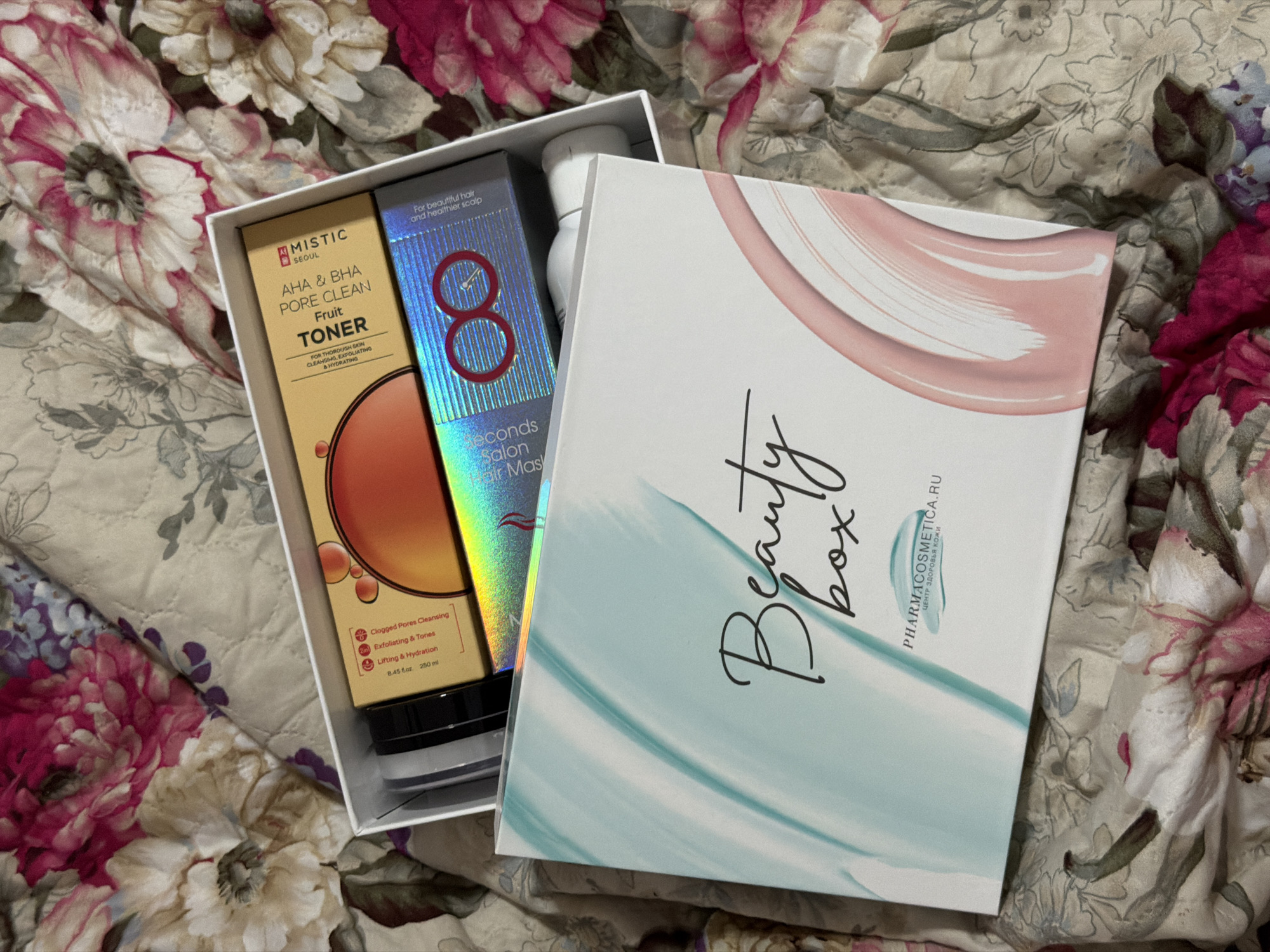 This is a top-down, medium shot photograph of an open beauty box on a quilted floral fabric. The beauty box is white with abstract teal and pink brush strokes on the lid. Inside the box are two skincare products. The first product, on the left, is a clear tube with a black cap and a black and white label. The second product, on the right, is a rectangular box with an orange gradient and a circular design. Text on this box reads "AHА & BHA PORE CLEAN TONER". Below this box is another product with a holographic, shimmering blue packaging. Text on this product reads "30 Seconds Salon Hair Mas". A white bottle with a white cap is visible in the bottom right of the box. The beauty box is open and positioned on a fabric with a large floral pattern in shades of pink, white, and muted green. The image does not contain any information about the city of Moscow, Russia.