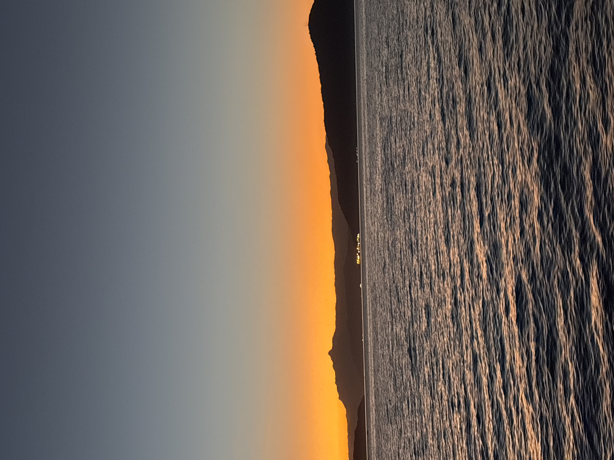 The image depicts a serene seascape at dusk, likely taken from a boat or the shore in Angra dos Reis, Brazil. The sky transitions from a deep, clear blue at the top to a vibrant orange hue near the horizon, indicating the sunset. Silhouetted against this colorful sky are several mountain ranges and hills. In the distance, lights from a town or settlement twinkle along the coastline. The foreground is dominated by the dark, rippling surface of the water, reflecting the warm tones of the sunset, creating streaks of orange and gold across the waves. The overall atmosphere is tranquil and picturesque, capturing the natural beauty of the Brazilian coast.