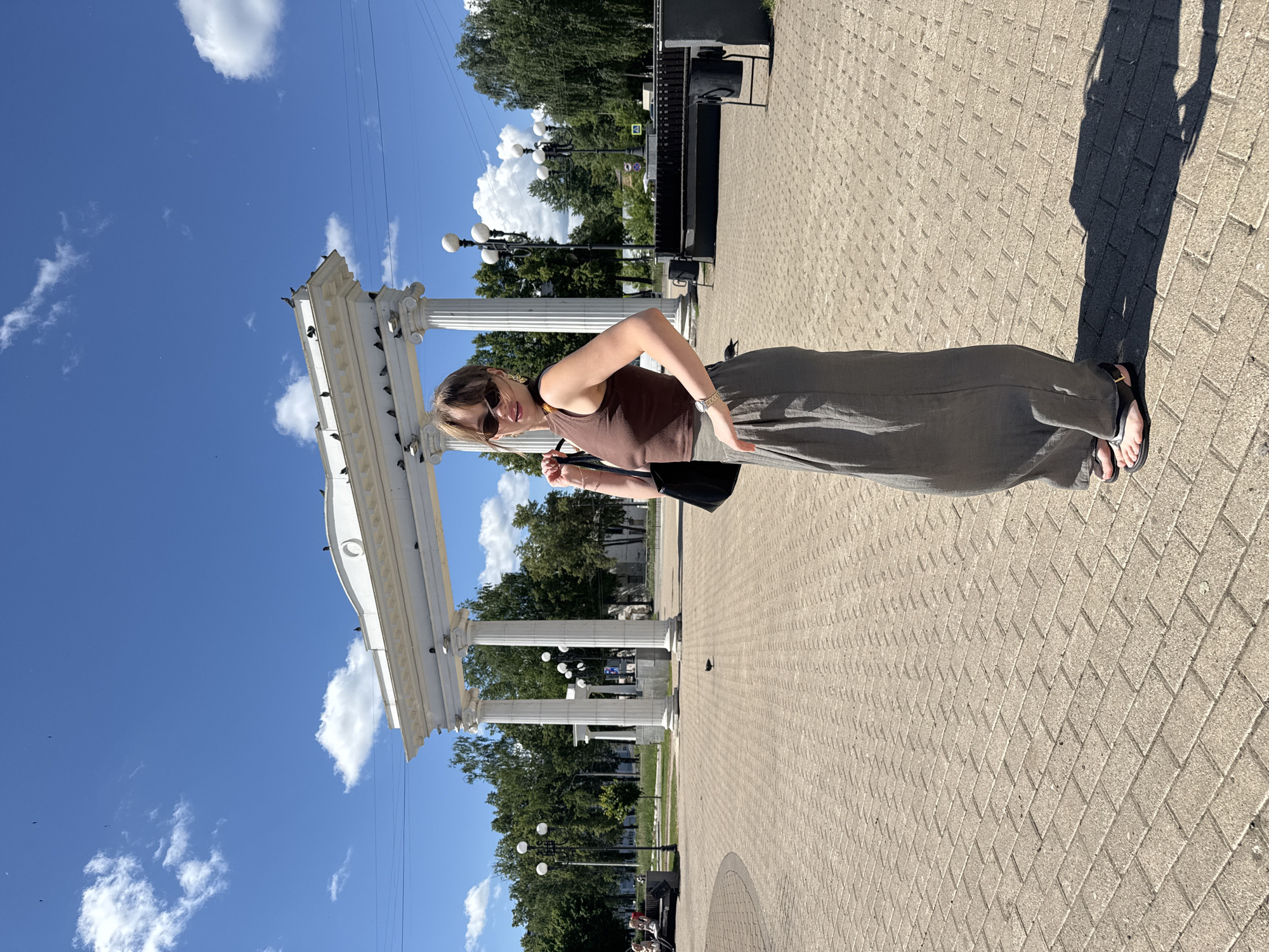 A full-body, profile shot shows a young woman with brown hair, sunglasses, and a brown tank top posing in a park in Chaikovsky, Russia. She is wearing loose, gray pants and black sandals. The woman is standing on a brick pathway in front of a white, neoclassical archway with columns. Trees and streetlights are visible in the background. The sky is blue with scattered white clouds.