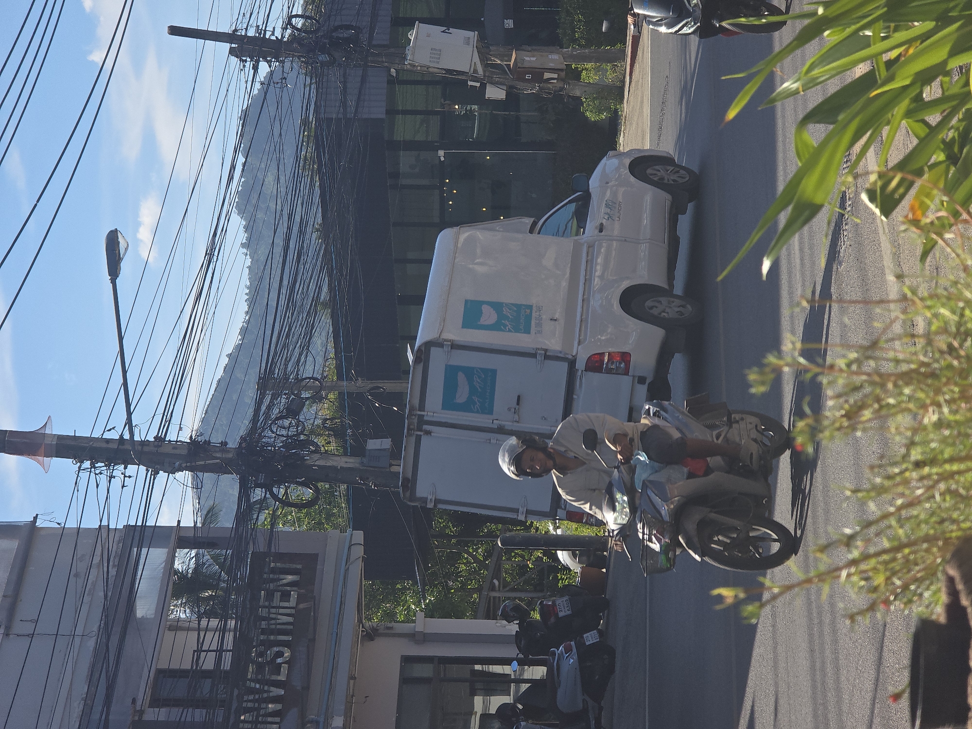 In this vertically oriented image, a street scene unfolds under a bright, partly cloudy sky, suggesting it's daytime. The foreground is dominated by lush green foliage, partially obscuring the view. A white van with "SA-ARD LAUNDRY" and a phone number printed on its side is parked on the right side of the road.
A person wearing a helmet is riding a scooter on the road, their face partially visible and their expression neutral. Behind them, more scooters are parked. In the background, a series of telephone poles with a dense network of wires stretches across the frame. A building with "INVESTMENT" clearly visible on its facade stands to the left, interspersed with electrical boxes and other urban infrastructure. The overall impression is a bustling street scene in an urban or semi-urban environment.