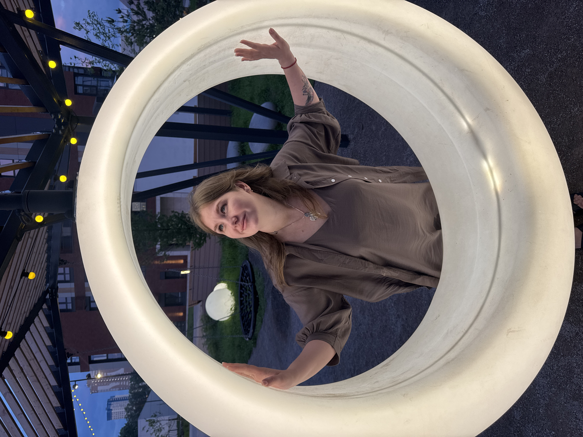 A young woman stands inside a large, circular, illuminated white frame. She is wearing a long-sleeved, brown button-down shirt and a necklace. She has her hands resting on the frame, with one arm raised in the air. The background includes buildings, greenery, and a pergola with small yellow lights. Based on the architectural details and the presence of a specific light fixture, the location appears to be in the city of Perm, Russia.