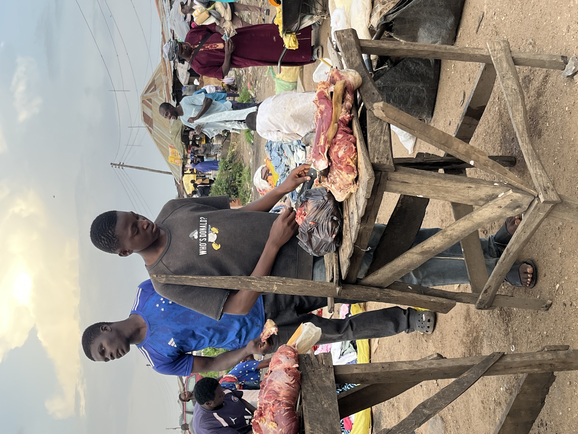 In Bukuru, Nigeria, an outdoor market scene unfolds under a cloudy sky. Two young men are prominently featured. The one in the foreground, wearing a dark t-shirt with the text "WHO'S DONALD?", is actively engaged in preparing meat. He stands behind a rustic wooden stall, holding a knife and interacting with a large cut of raw meat on the counter. To his left, another young man in a blue t-shirt watches, holding a piece of meat in his hand.
Surrounding them, the market buzzes with activity. Numerous people mill about, some dressed in traditional attire like the woman in a maroon robe holding money. Stalls are set up, some covered with corrugated metal roofs, displaying various goods. Piles of fabric and sacks are visible, and a wheelbarrow is present. The ground is dusty, indicating a dry, open-air environment. The overall atmosphere suggests a vibrant, everyday market in an urban setting.