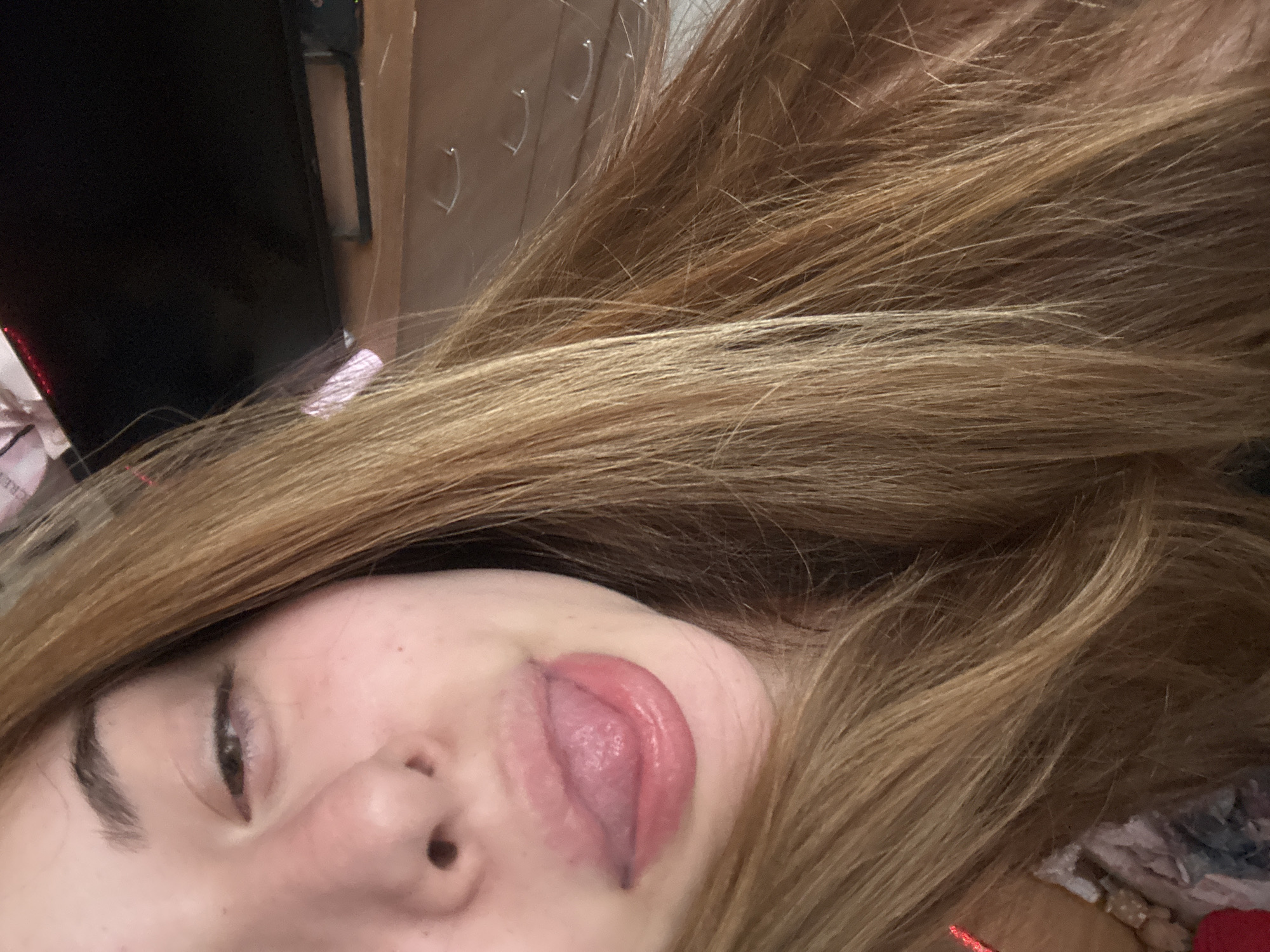 This close-up image, likely a selfie, features a person with long, light brown to blonde hair dominating the right side and foreground. The individual's face is partially visible, showing the left eye with a dark eyebrow, a portion of the nose, and the mouth. The person is engaged in a playful act, sticking their tongue out, which is prominently displayed in the lower central part of the frame. The tongue is pinkish-red with some darker lines.
The background elements are somewhat indistinct but suggest an indoor setting. To the upper left, a large dark rectangular object, possibly a turned-off monitor or television screen, is partially visible. Above the person's head and towards the right edge, a lighter brown surface appears, adorned with several small, white, hook-like clips or hangers arranged horizontally. Other blurry shapes and textures in the background indicate general indoor clutter. The lighting is dim and artificial, consistent with an interior shot, possibly taken during the evening. The image conveys a lighthearted and informal mood. No visible text is present in the image. The provided location context is Troyan, Bulgaria, but no specific visual cues within the image confirm this.