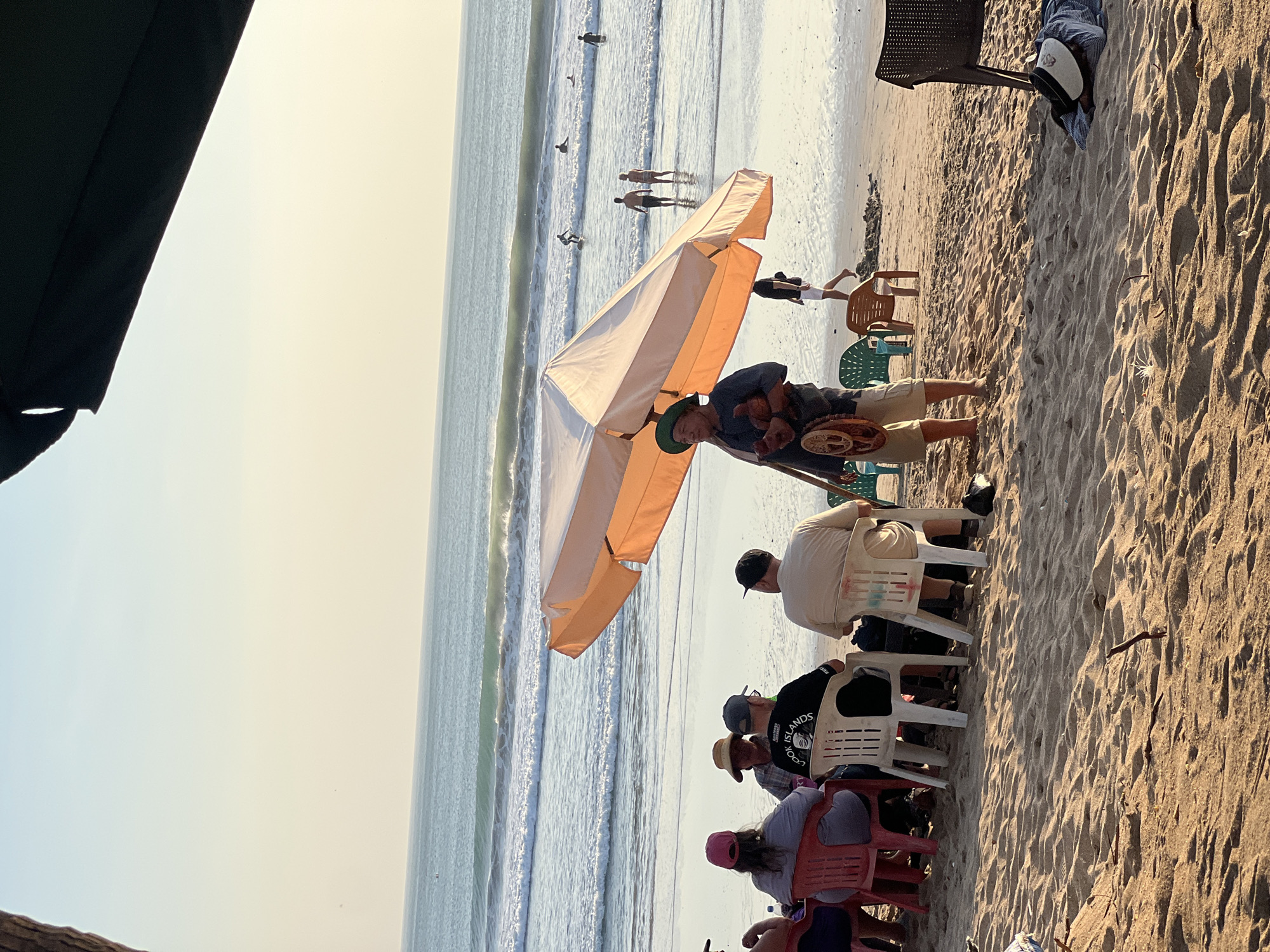 The image depicts a beach scene in Kuta, Indonesia, likely during the late afternoon given the warm, soft lighting and the color of the sky. In the foreground, a group of people are seated at tables on the sand, under an orange umbrella. They are facing away from the camera, towards the ocean, and appear to be enjoying a meal or drinks. One person in the group is wearing a hat that says "Cook Islands".
A man standing under the umbrella is playing a stringed instrument and holding a wooden mask. He is wearing a hat and shorts. Further back, the ocean stretches out, with gentle waves breaking on the shore. Several people are in the water, some surfing and others wading. The beach is sandy, with some scattered beach chairs and debris. The overall atmosphere is relaxed and leisurely, characteristic of a tropical beach destination.