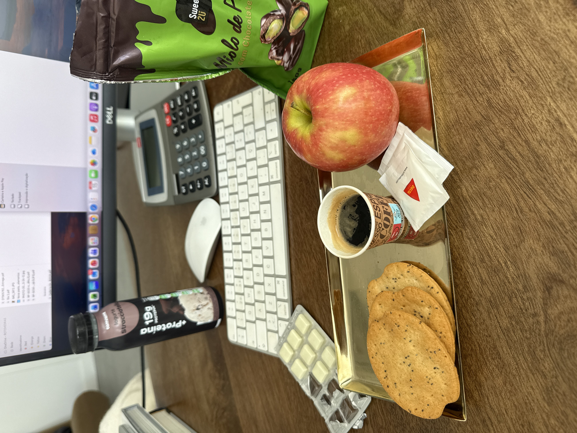 On a wooden desk, a collection of items suggests a workspace snack break. A monitor displaying files, a calculator, and a white wireless keyboard and mouse indicate a computer workstation. A golden tray holds an apple, coffee, two sugar packets, and three crackers. Adjacent to the tray is a chocolate protein shake, a partially consumed white and dark chocolate bar, and a bag of chocolate-covered pistachios labeled "Miolo de Pistachio com Chocolate," indicating a location in Portugal, specifically the city of Torres Vedras.