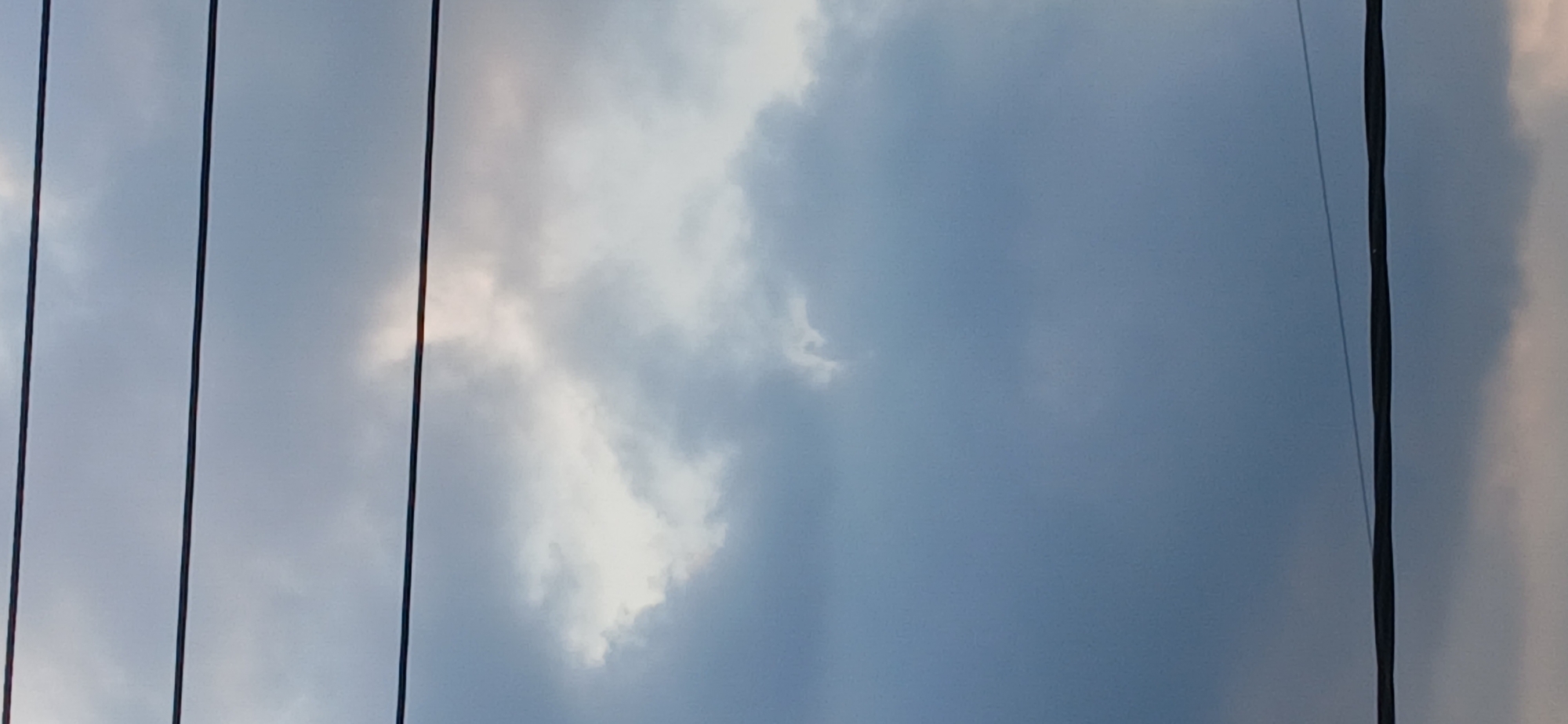 The image depicts a view of the sky through multiple power lines, likely in Tenggarang, Indonesia. The sky is predominantly filled with overcast, gray clouds, with a sliver of brighter sky and possibly sunlight visible in the upper central portion of the frame. There are three distinct dark, horizontal power lines stretching across the image. The lower part of the image shows a band of clouds with a subtle pinkish hue, suggesting either a sunrise or sunset. There are no people, animals, or specific structures visible in the frame, making it a purely environmental shot focusing on the sky and overhead wires.