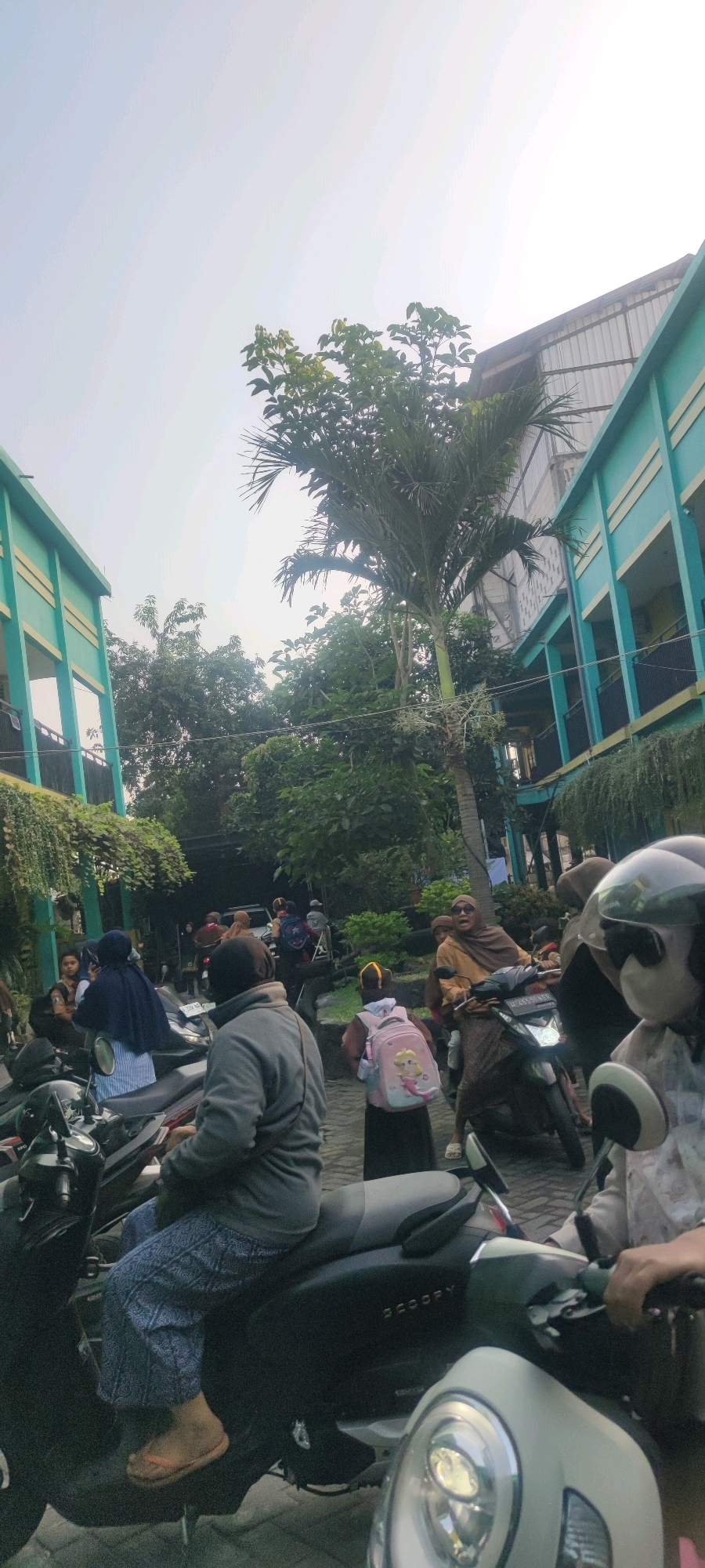 In Banjarsari, Indonesia, a group of people are gathered outdoors in front of a turquoise building with multiple floors, possibly a school or an educational institution. The sky is bright and overcast. Several motorcycles are parked, and some individuals are seated on them. One person in the foreground is wearing a grey jacket and patterned pants, with an orange flip-flop on their left foot. To their right, another individual wearing a light-colored shirt and sunglasses is partially visible, seated on a white scooter. A young person with a pink backpack featuring a cartoon character is walking away from the camera. To their left, a person wearing a dark blue hijab is seated on a motorcycle. Further back, a woman in a brown hijab is riding a motorcycle and looking towards the camera with her mouth open. The environment is lush with trees and foliage.