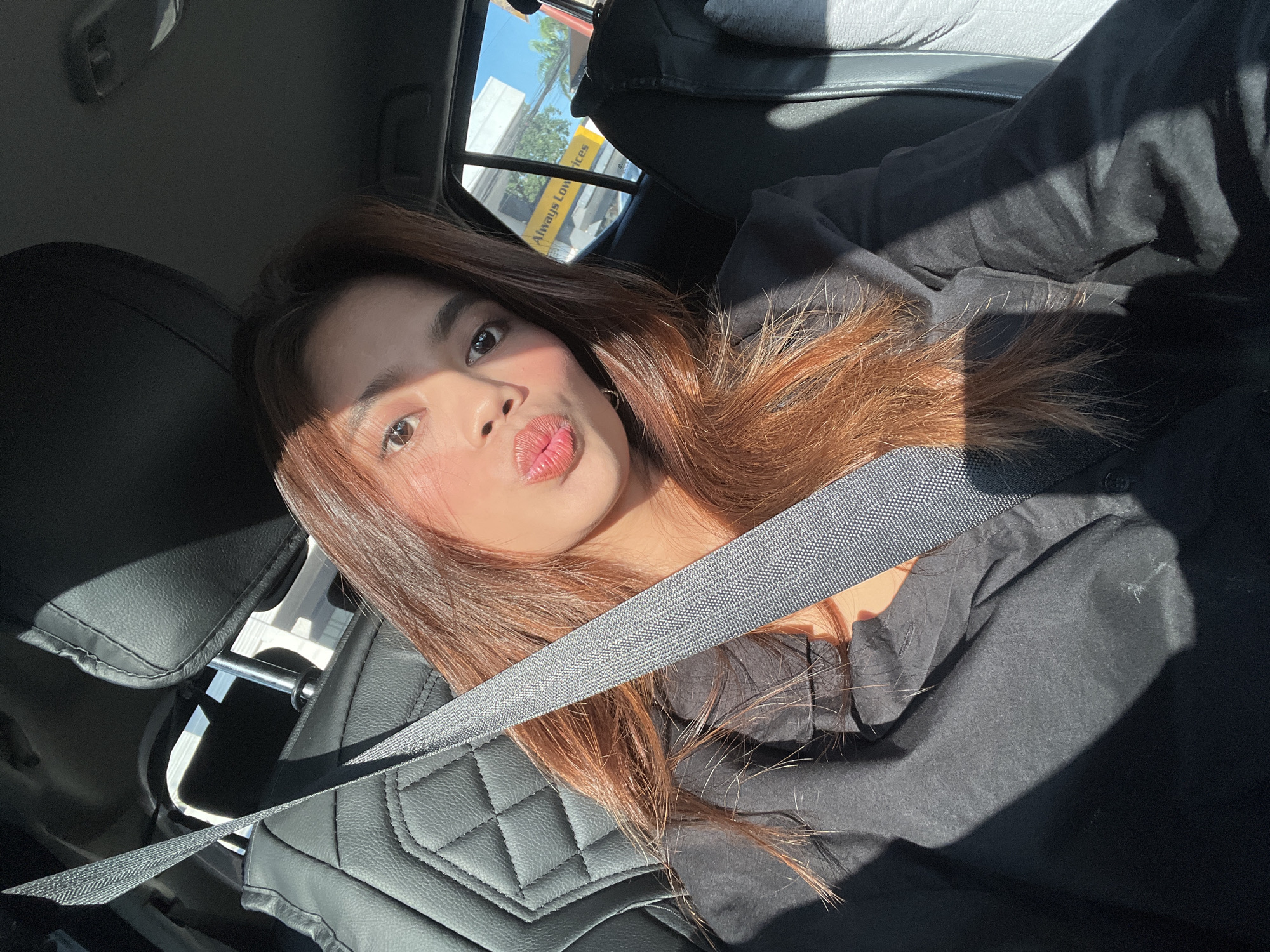 The image captures a selfie of a young woman taken inside a vehicle. The woman has long, reddish-brown hair and is wearing a black long-sleeved shirt, with a gray seatbelt positioned diagonally across her chest and shoulder. She is pouting with puckered lips. Strong sunlight illuminates the right side of her face and hair, casting a distinct shadow across her left shoulder and torso. The vehicle's interior features dark-colored, possibly leather, seating with a quilted or diamond-stitched pattern visible on the seatback behind her.
Through the rear passenger window, an external scene is visible under clear, bright skies. A prominent bright yellow commercial sign with black lettering is a key feature. The text "Always Low Prices" is clearly legible on this sign, which is the signature slogan of Puregold Price Club, a major supermarket chain in the Philippines. Green foliage and parts of a building are also visible in the background, characteristic of an urban or commercial environment. Based on the presence of the "Always Low Prices" sign, the scene is located near a Puregold supermarket branch within a commercial area of Davao City, Philippines.