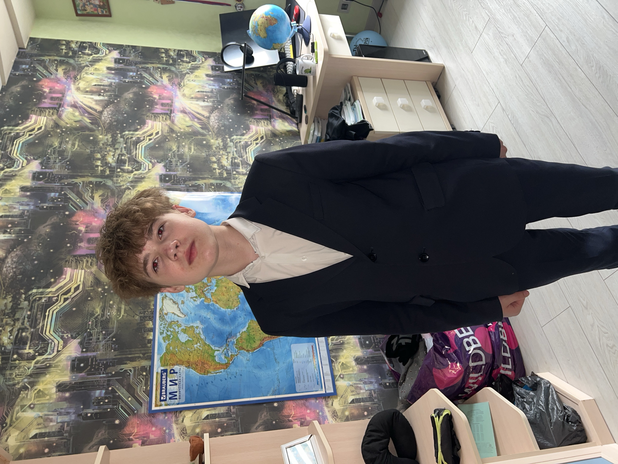 A fair-skinned young man with curly, light brown hair is standing indoors, wearing a dark blue suit jacket over a white button-down shirt. The background consists of a wallpaper with a futuristic circuit board design, a world map poster with the Russian word "МИР" (World) visible, and a desk. On the desk, there is a globe, a monitor, and various items. To the right, there are some bags and organized bins. The location cannot be determined to be in Perm, Russia with the given details.