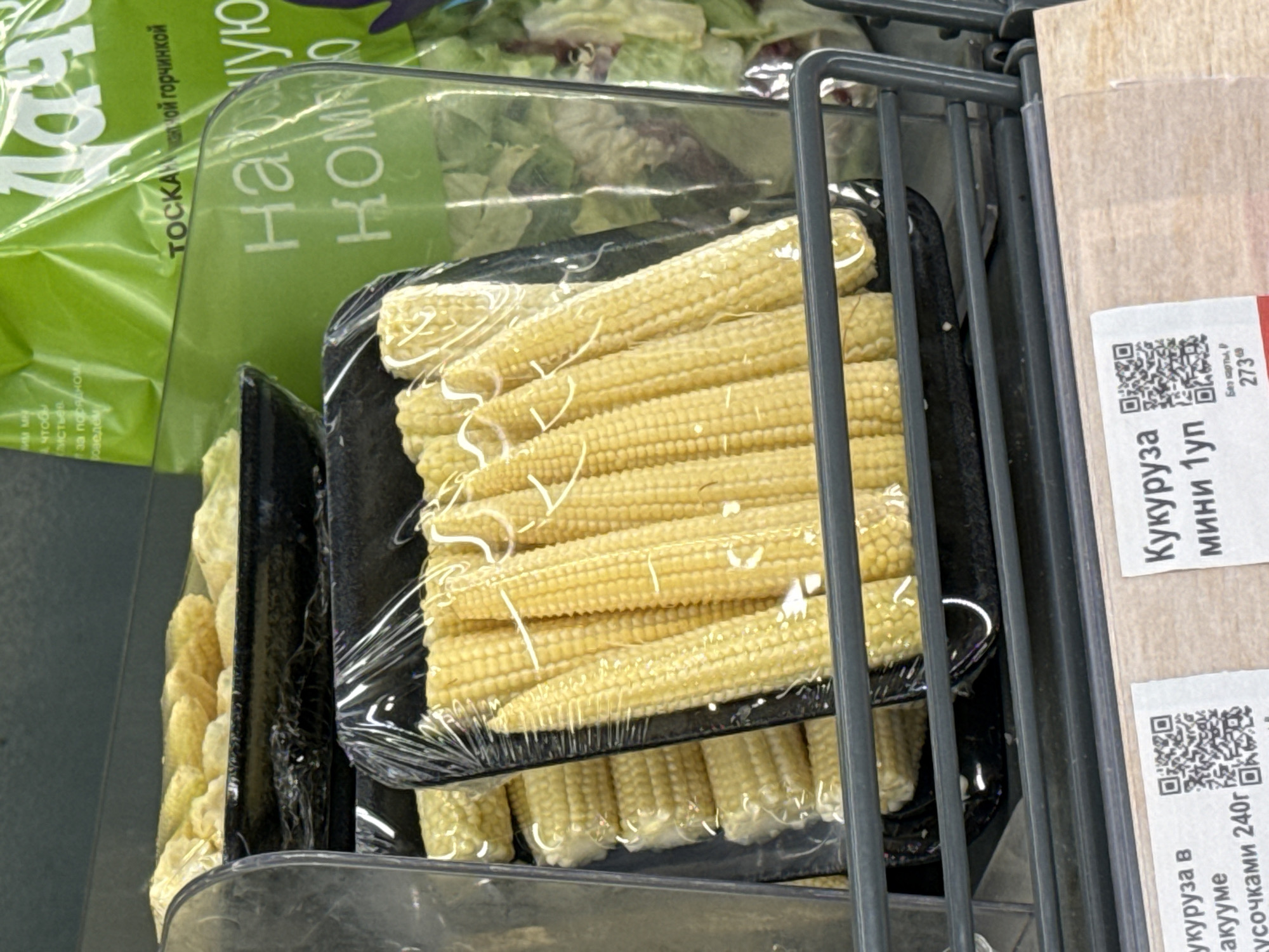 The image shows packages of baby corn in a shopping cart, likely in a supermarket in Izhevsk, Russia. The corn is arranged in black plastic trays, covered in clear plastic wrap. A label on the right side of the image reads "Кукуруза мини 1уп" (Corn mini 1 pack) and "кукуруза в вакууме кусочками 240г" (Corn in vacuum in pieces 240g), along with a QR code. To the left of the corn, there is a bag of salad and part of another package of baby corn. The shopping cart is grey metal.